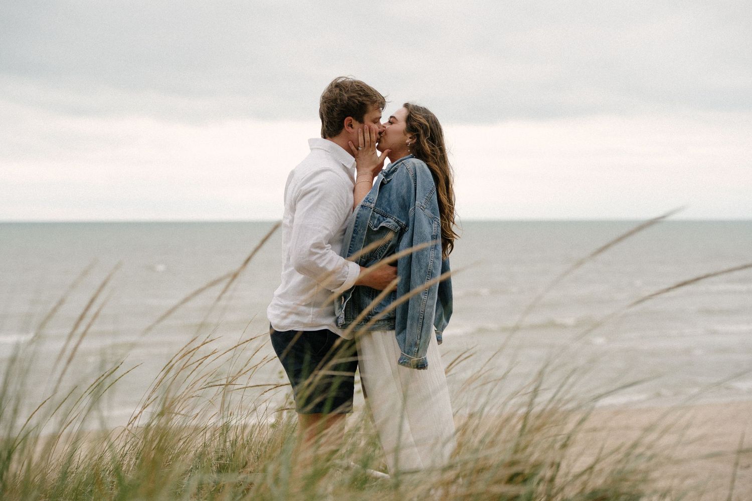 Couple sharing a romantic kiss among tall beach grass with the ocean in the background on a cloudy day.