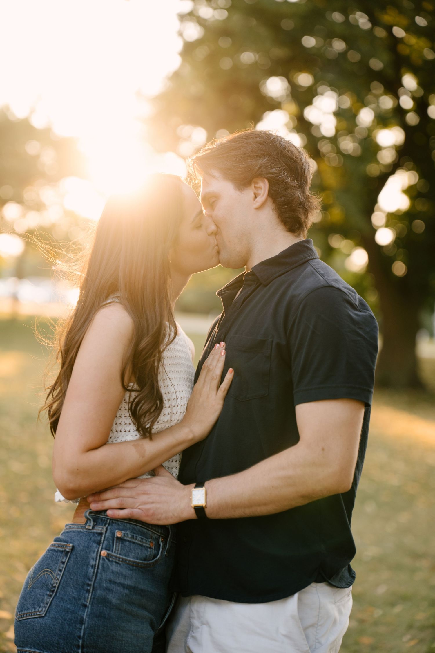 Couple kissing in a sun-drenched park, woman in jeans, man in black polo shirt.