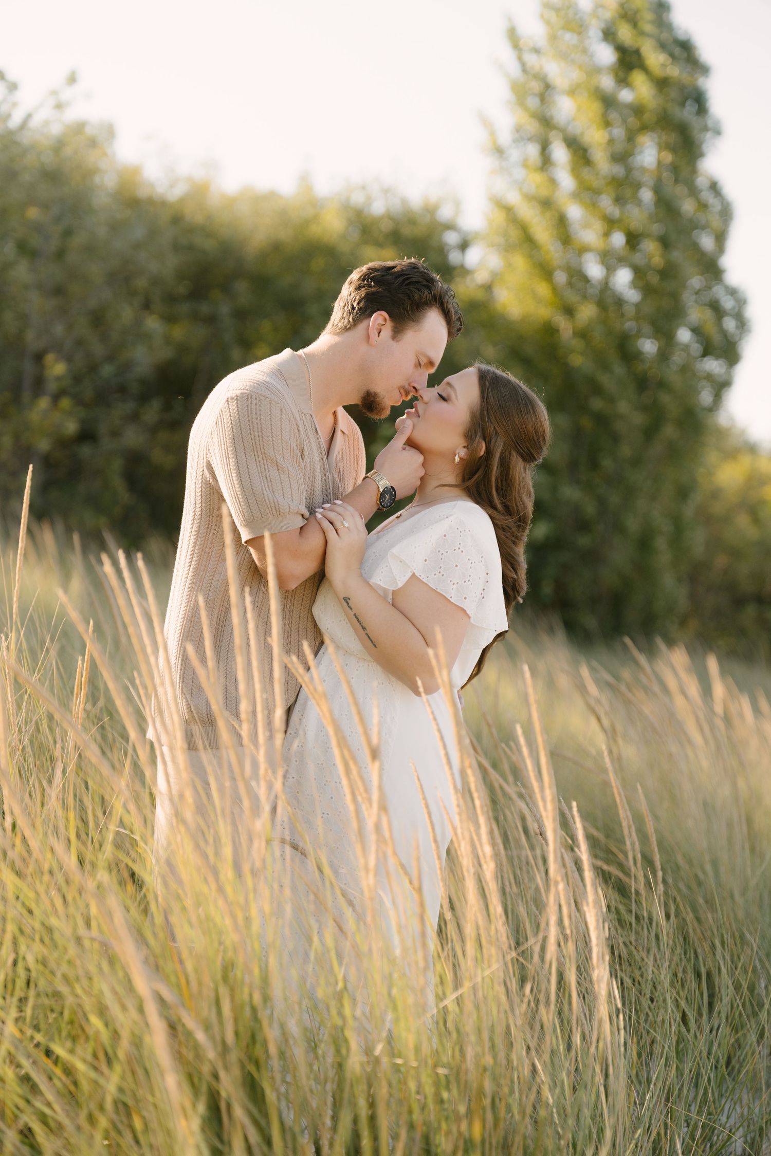 Couple embracing and kissing in tall golden grass field during sunset engagement session.