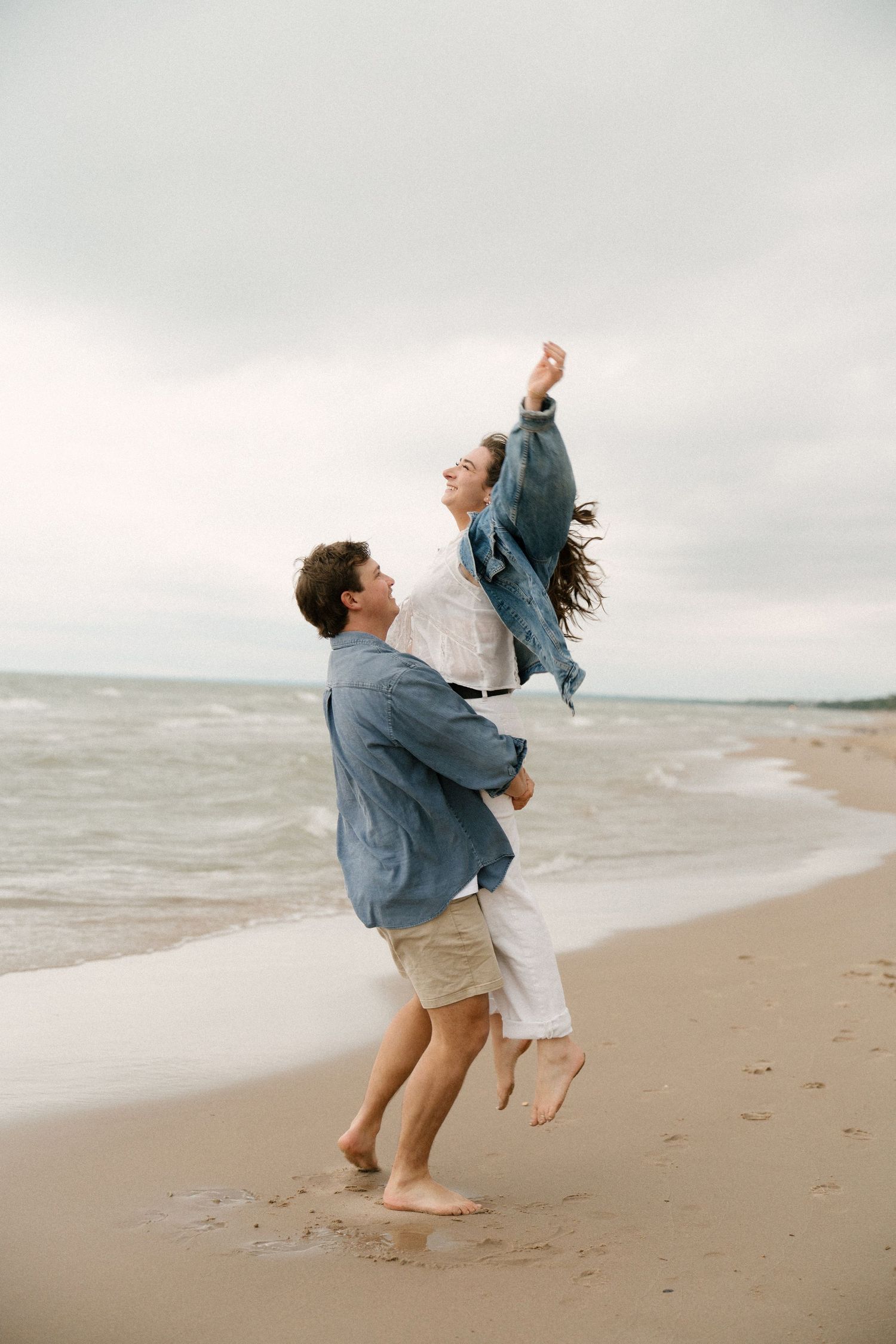 Joyful man lifting his laughing girlfriend on a sandy beach, both wearing casual outfits with ocean waves in background.