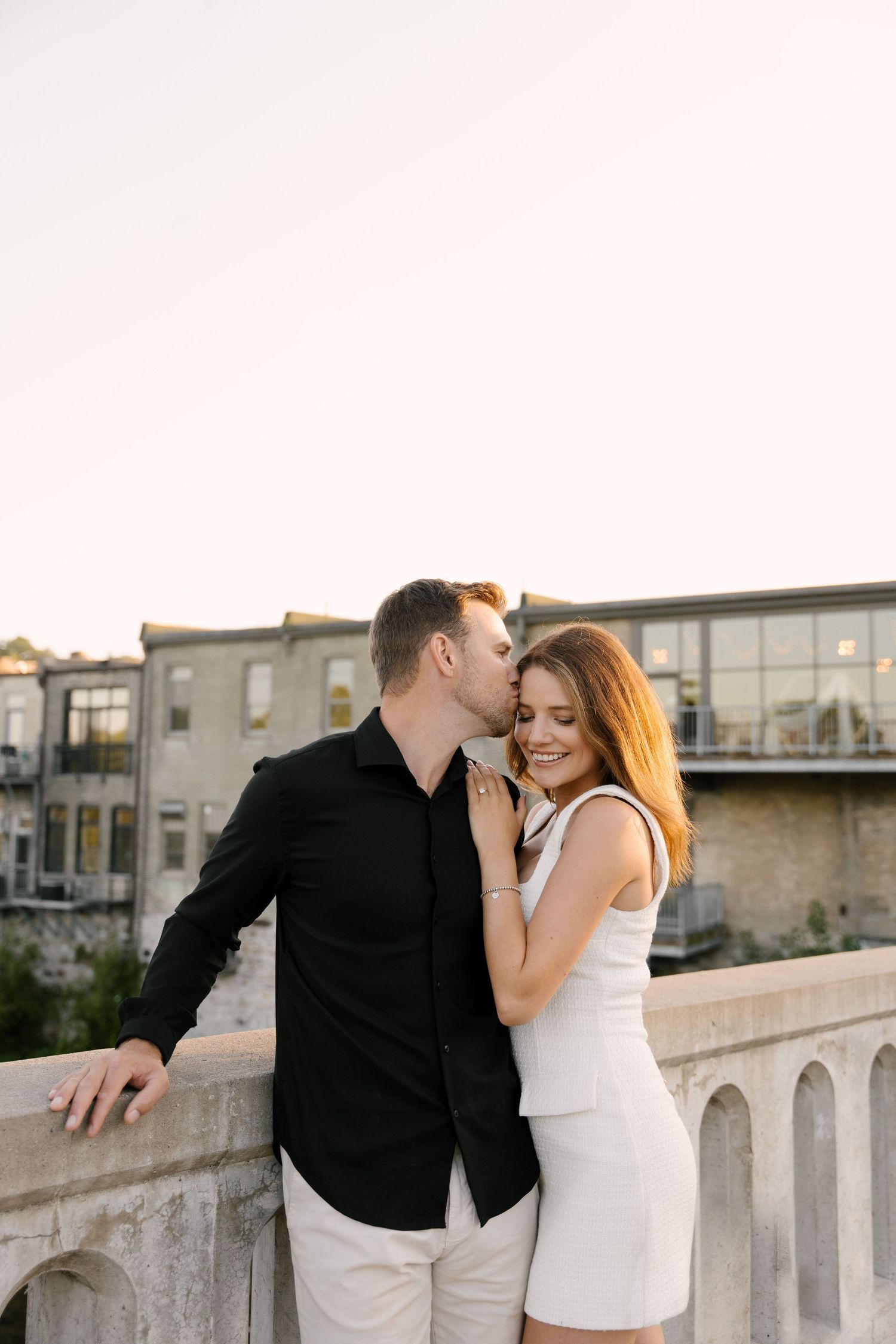 Couple sharing a romantic kiss on a stone bridge at sunset with industrial loft buildings in the background.