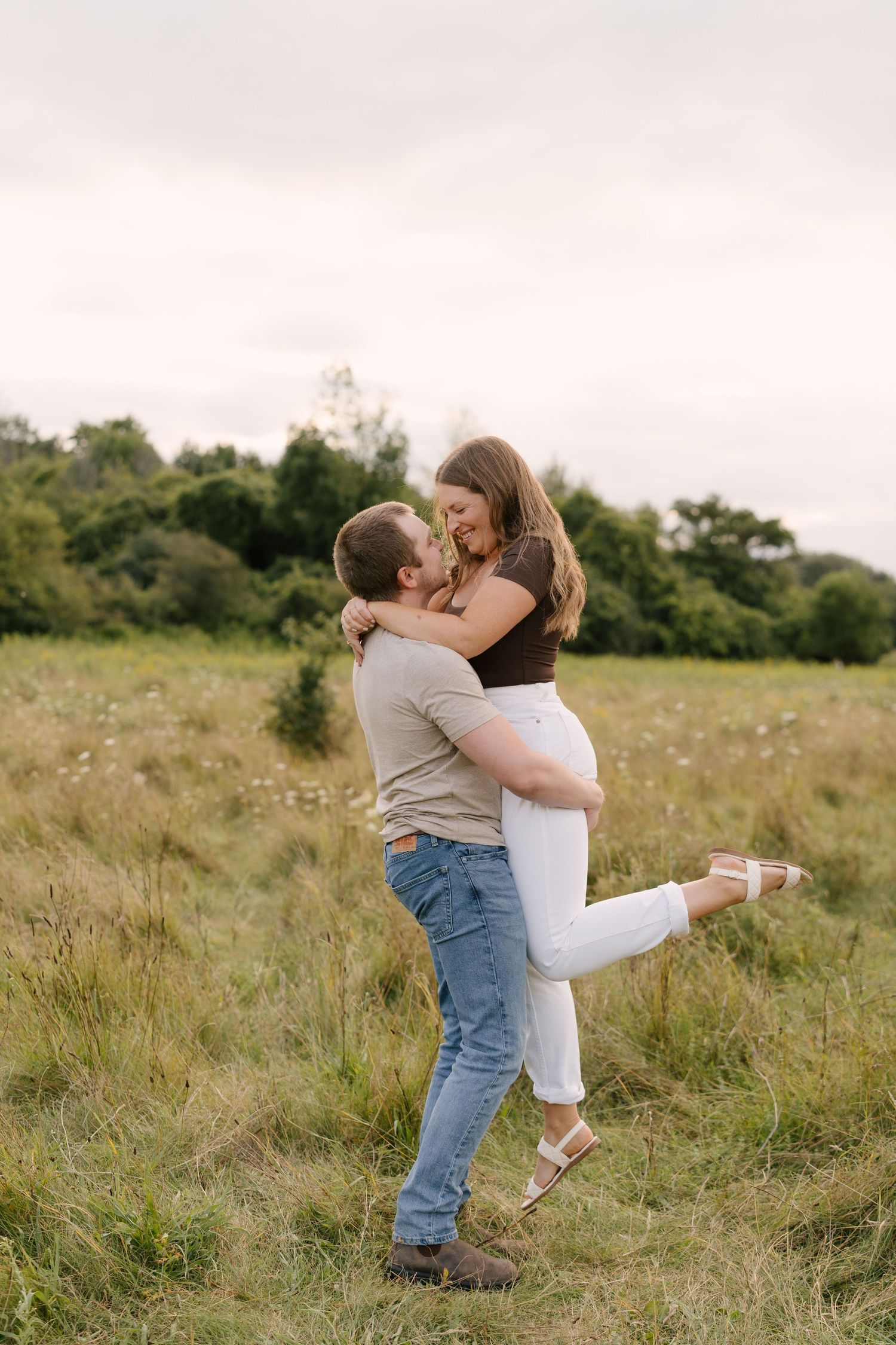 Man lifting woman in a joyful embrace in an open meadow with green trees in the background.