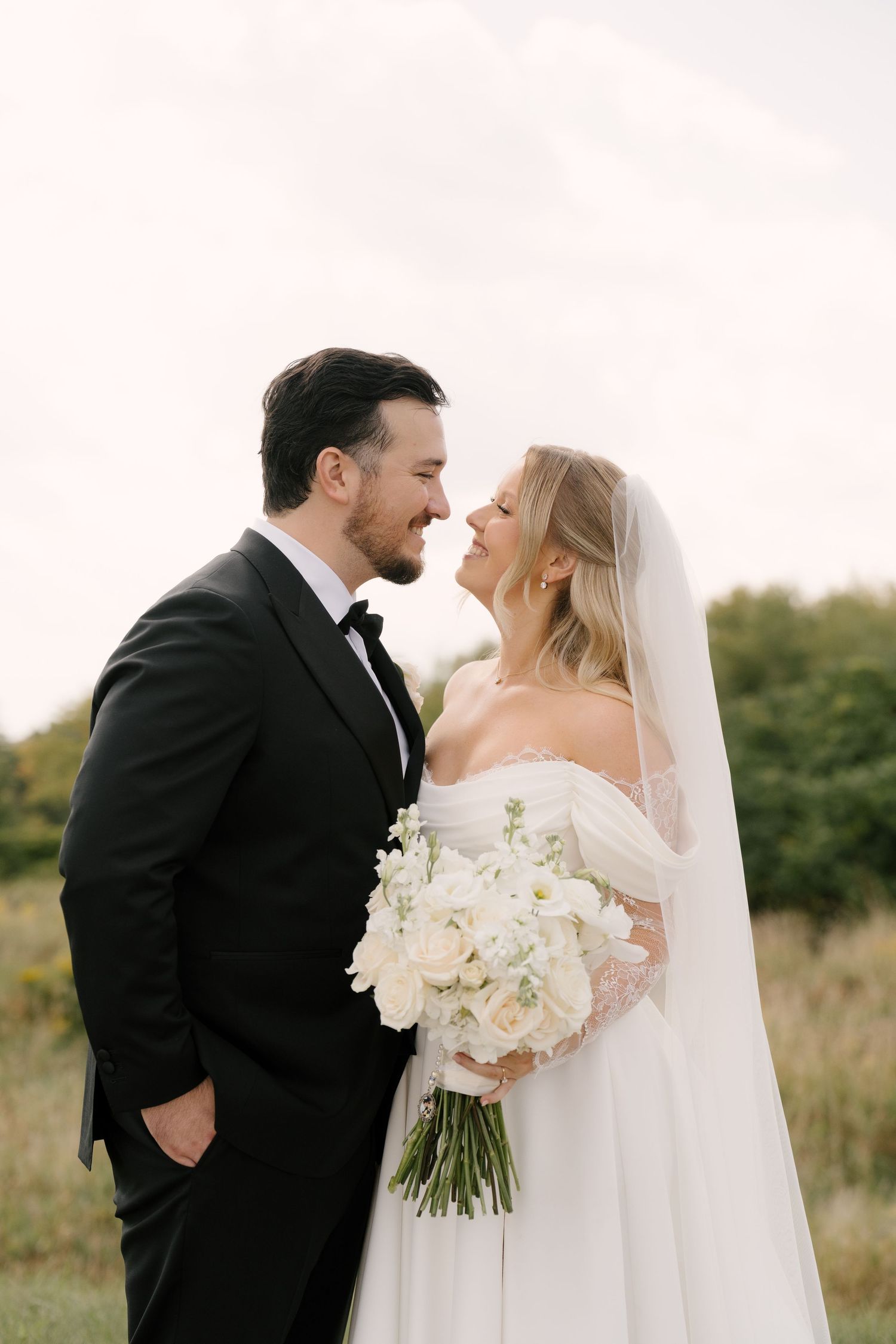 Bearded groom in black tuxedo and blonde bride in off-shoulder gown share intimate moment outdoors.
