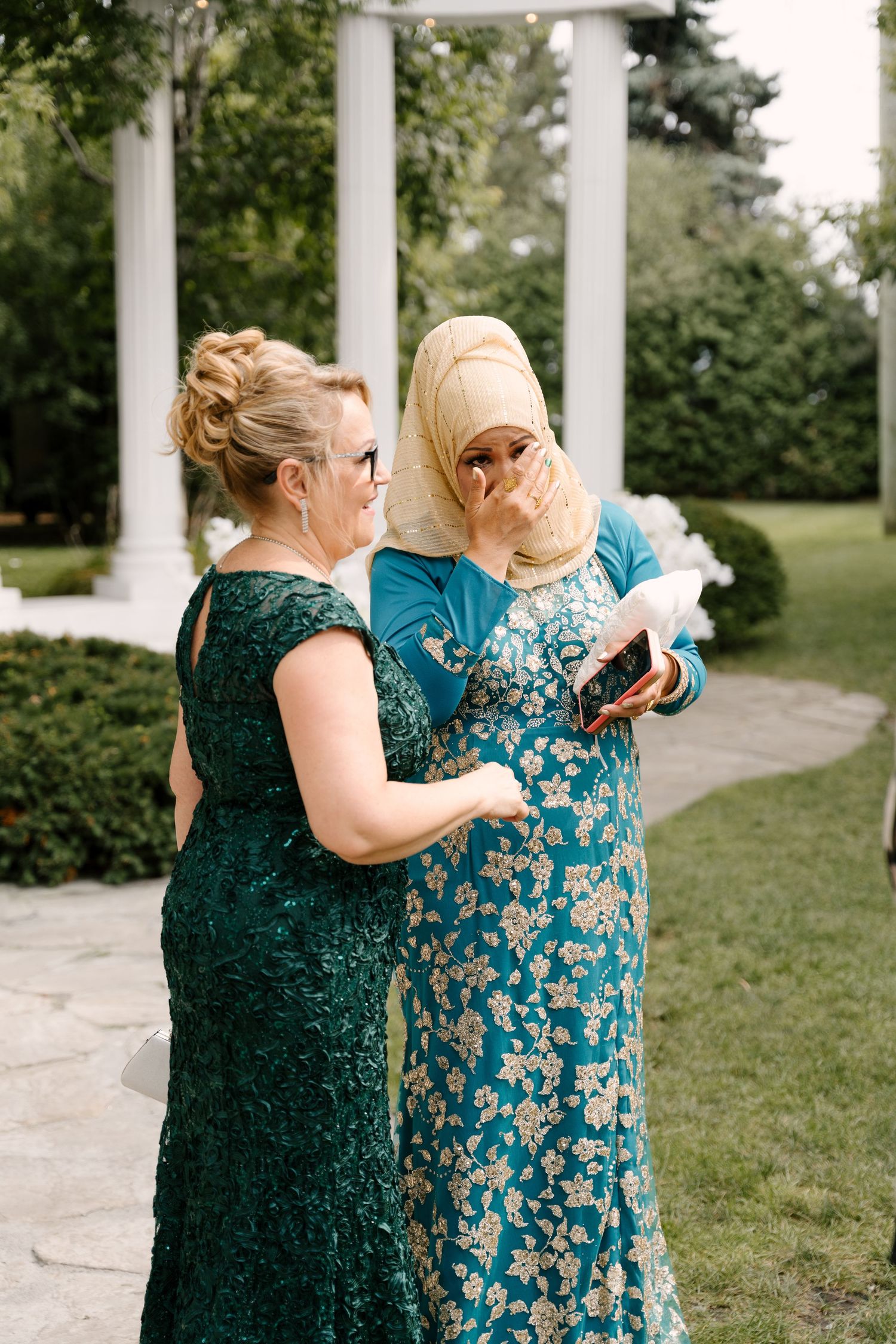 Two emotional female wedding guests in teal dresses view photos on phone at outdoor garden ceremony.