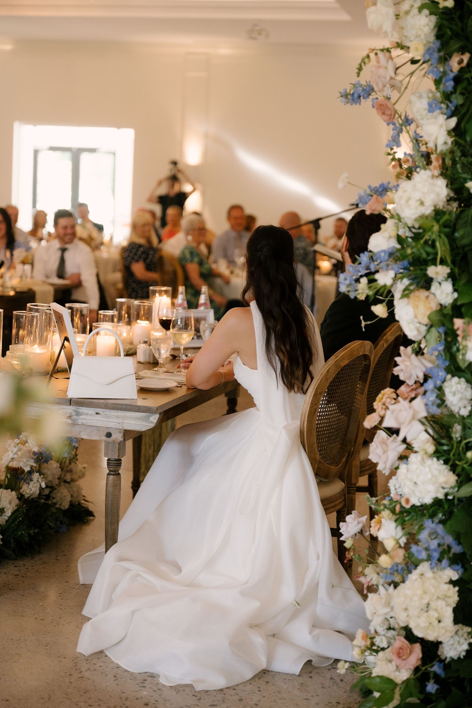 Bride in white gown seated at sweetheart table surrounded by floral arrangements at Roseville Estates wedding reception.