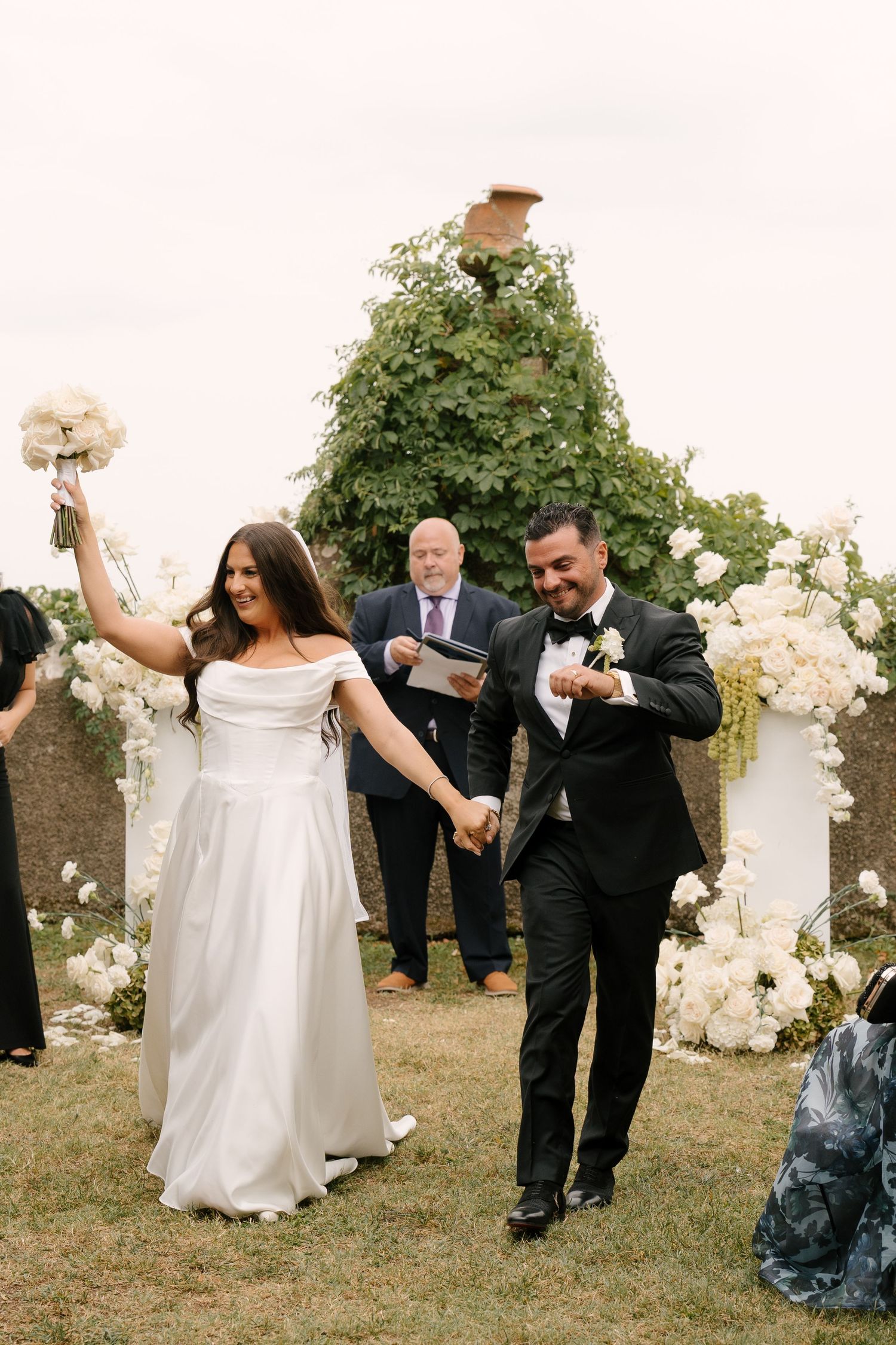 Joyful newlyweds walk back down aisle after Tuscan wedding ceremony, bride raising bouquet in celebration outdoors.