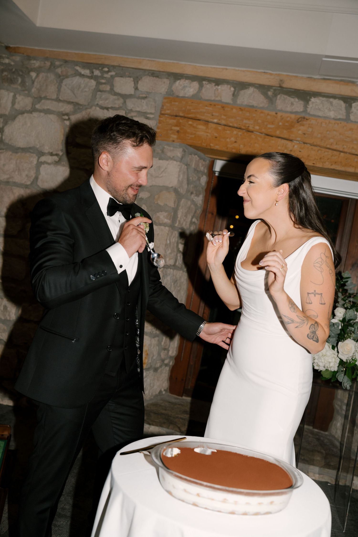 Bride and groom in black tuxedo feeding each other wedding cake near stone fireplace at reception in Cambridge Mill.