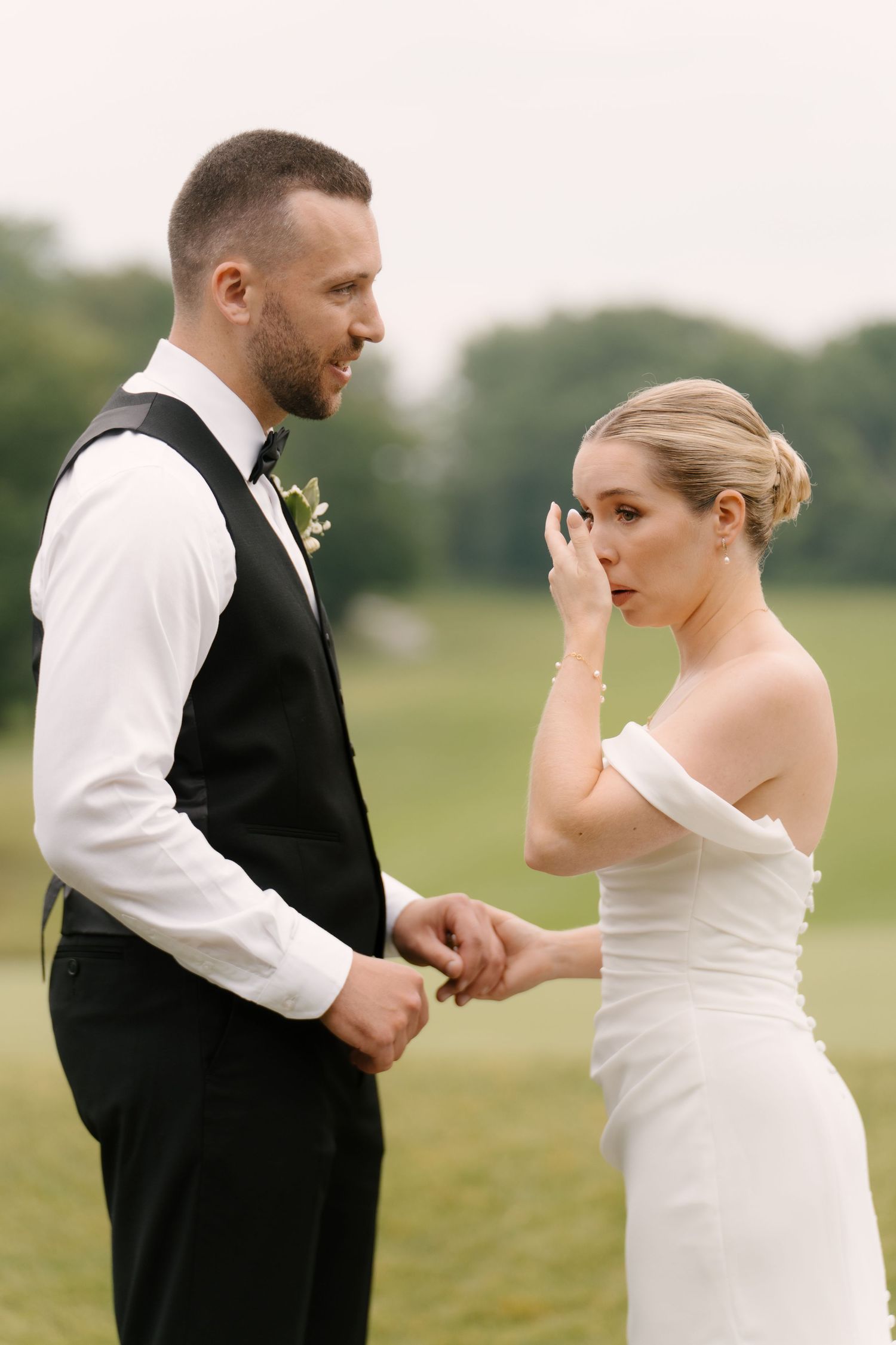 Emotional bride wiping tears while groom holds her hands during outdoor vow exchange.