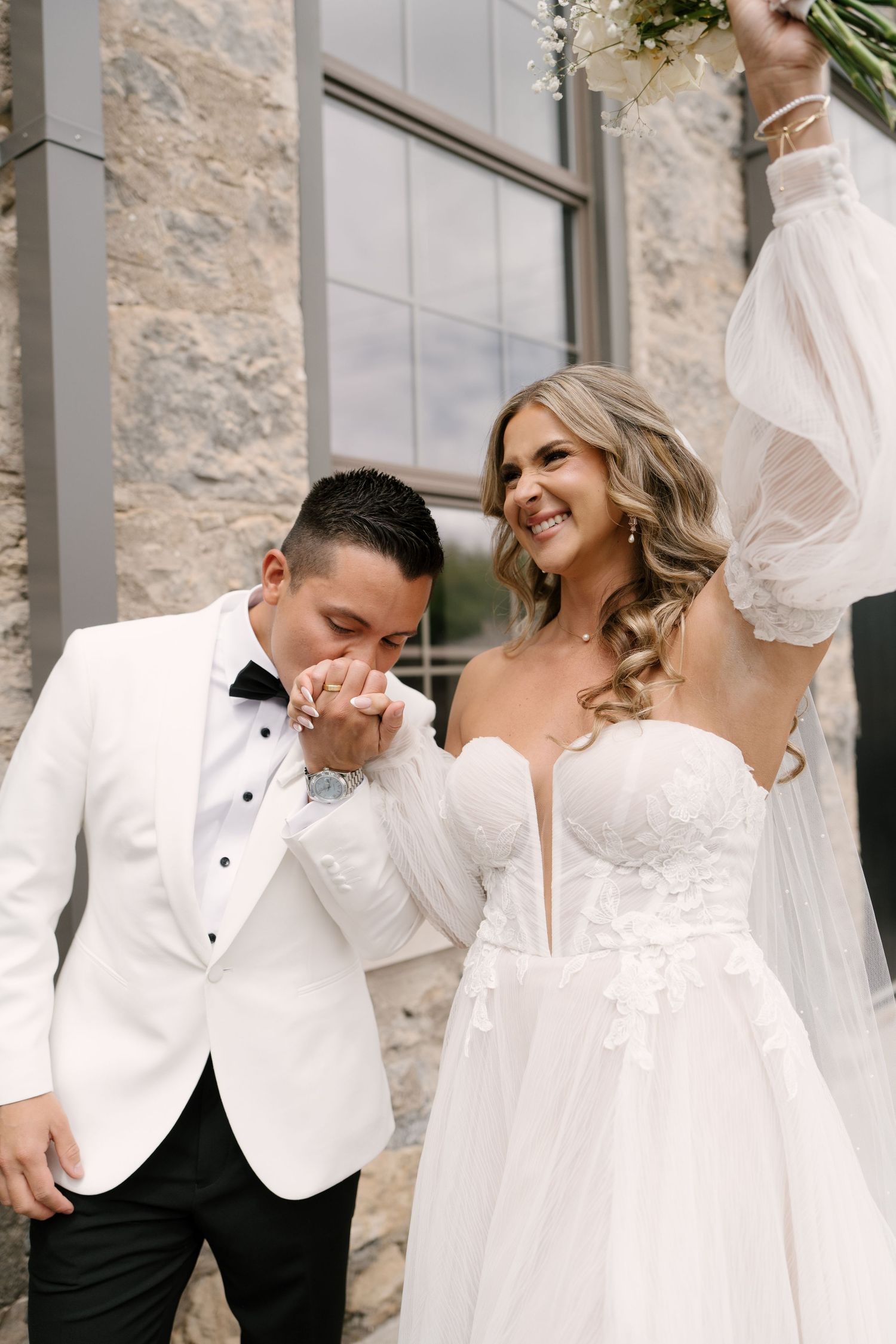 Groom in white tuxedo kissing bride's hand as she smiles and raises bouquet at outdoor wedding at Tapestry Hall wedding venue.
