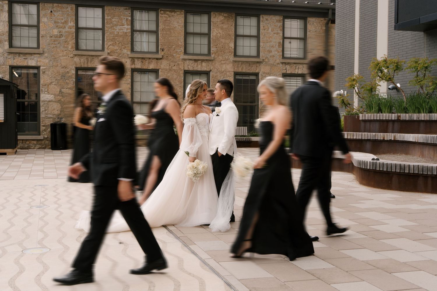 Bride and groom share a kiss surrounded by wedding party walking in motion blur outdoors at Tapestry Hall wedding venue.