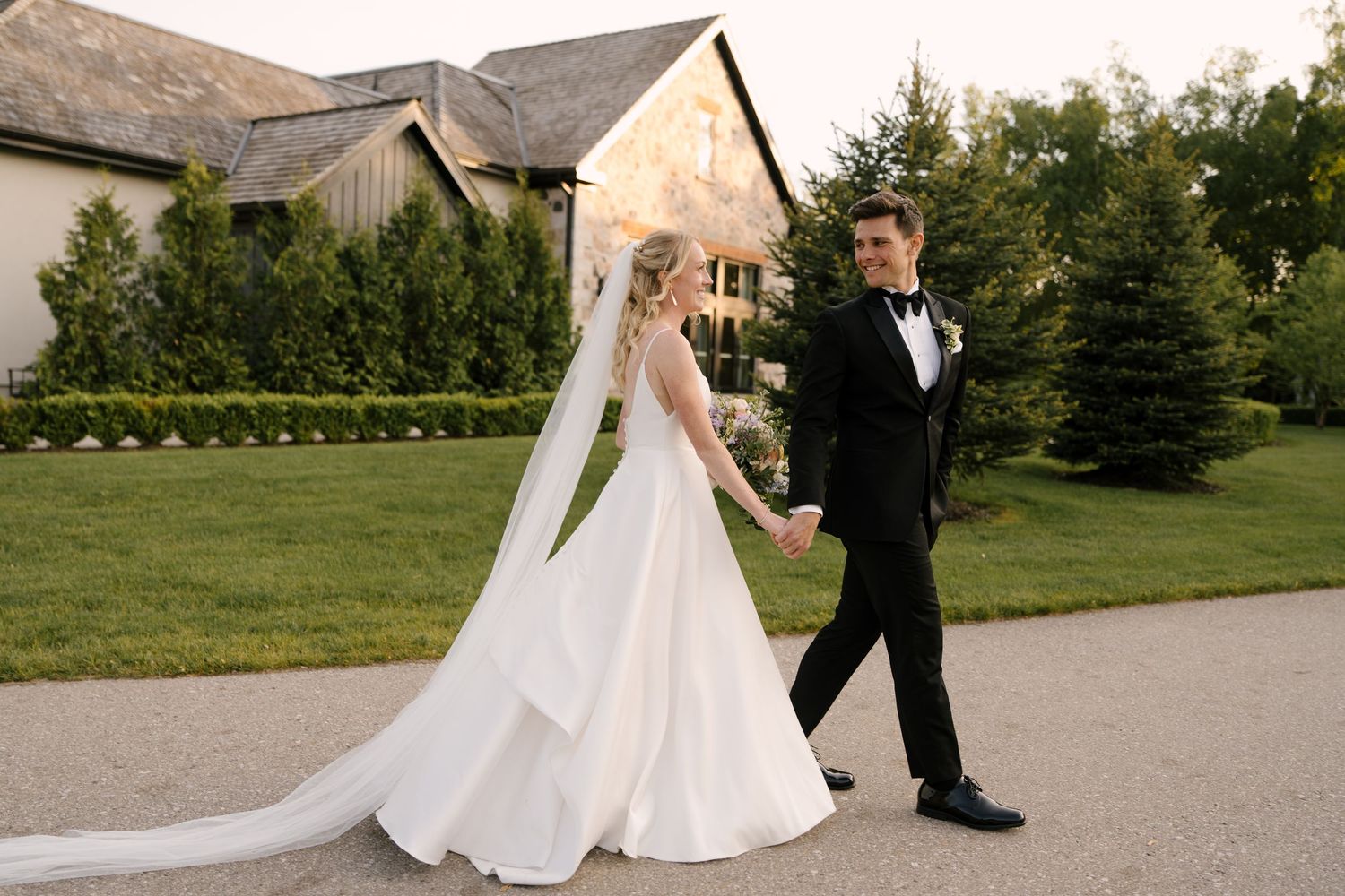 Bride in white gown and groom in black tuxedo walk hand in hand outdoors at elegant Roseville Estates wedding venue.