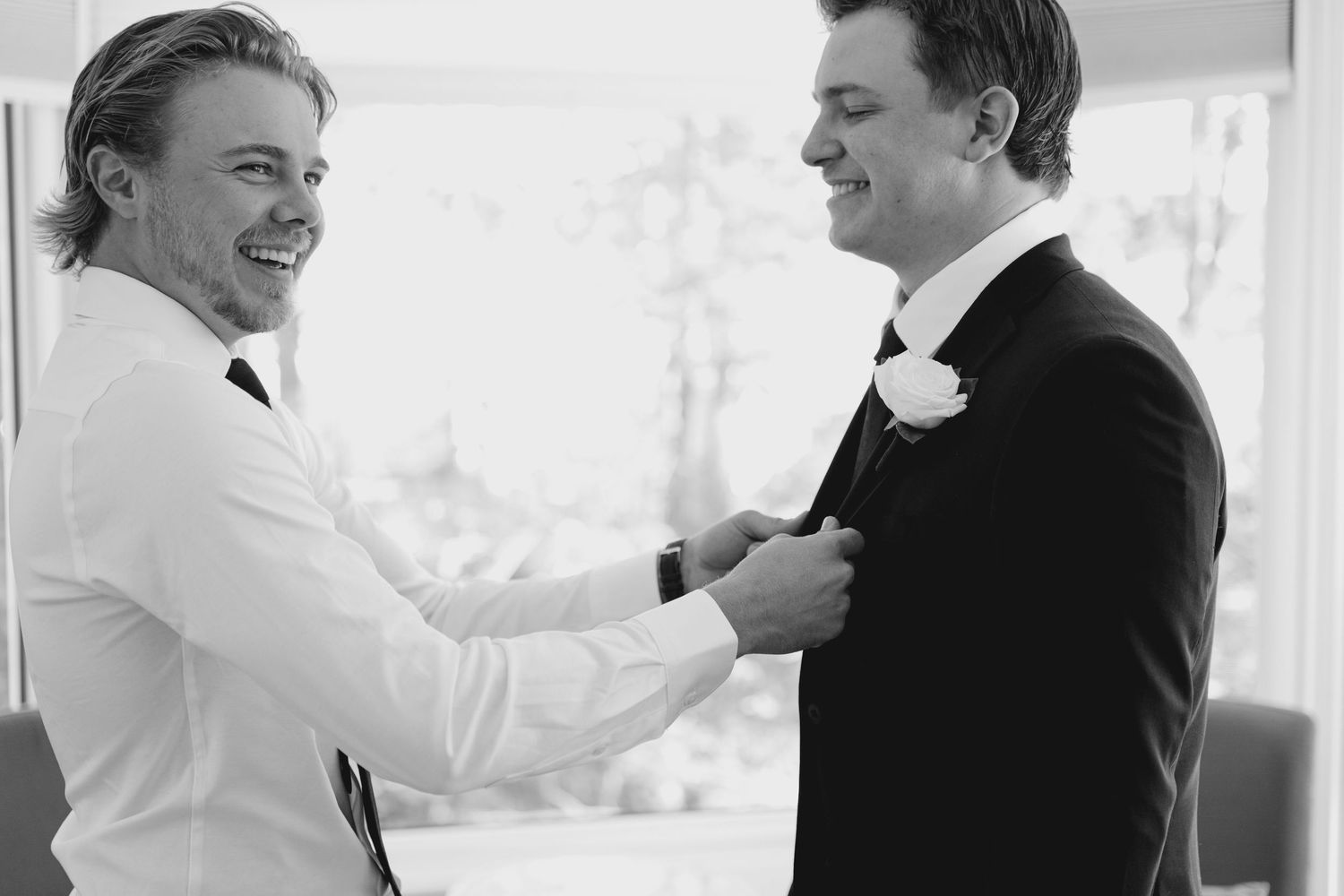 Groomsman in white shirt adjusts boutonniere on groom in black tuxedo, black and white photo.