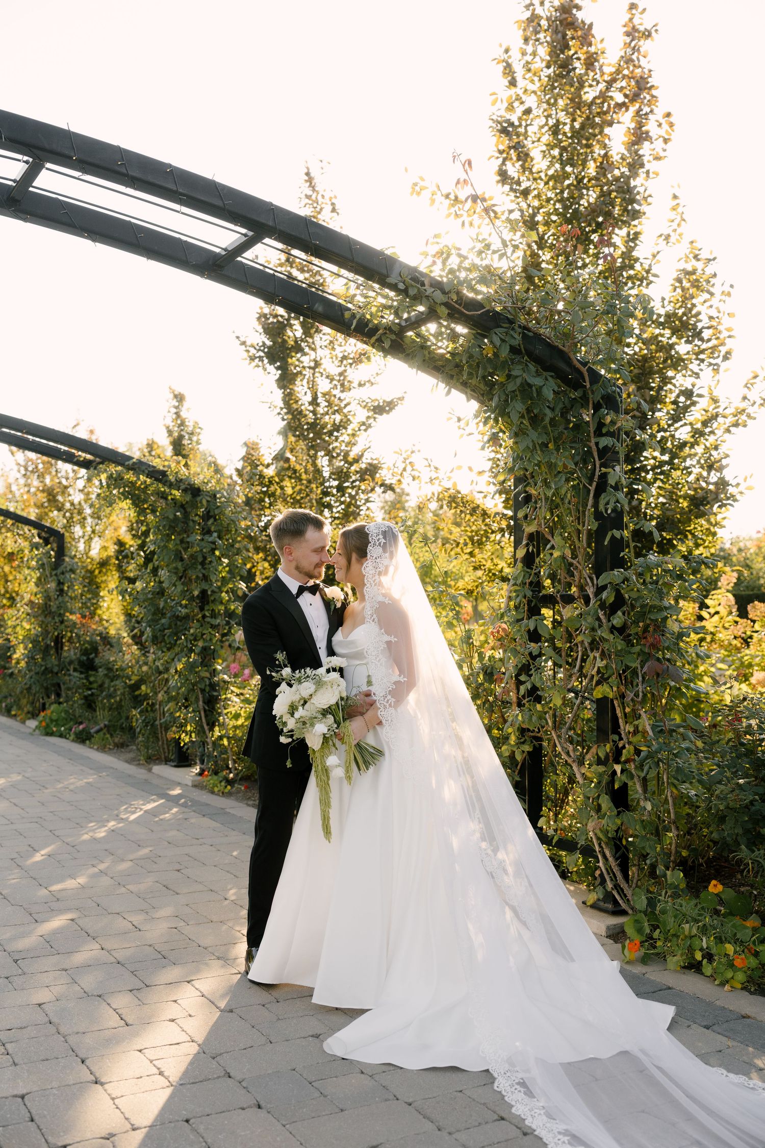 Bride and groom share a romantic moment under a lush garden arch, bride holding white bouquet with flowing veil at Pillar and Post.