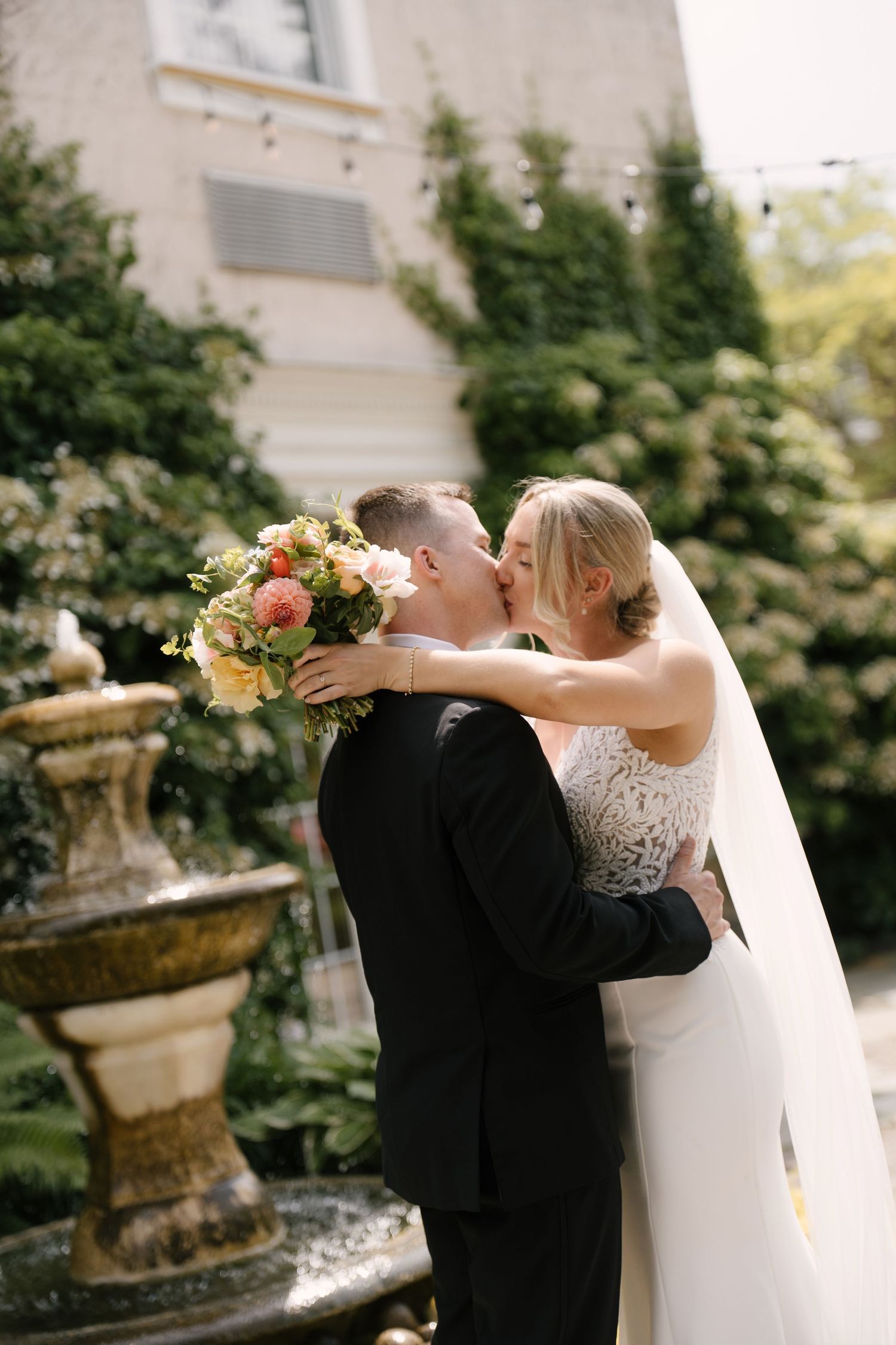 Bride and groom share passionate kiss beside garden fountain holding colourful bridal bouquet at Inn On The Twenty.