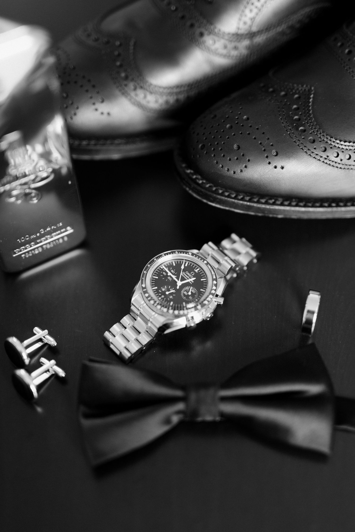 Black and white flat lay of groom accessories including watch, bow tie, cufflinks, cologne and oxford dress shoes.
