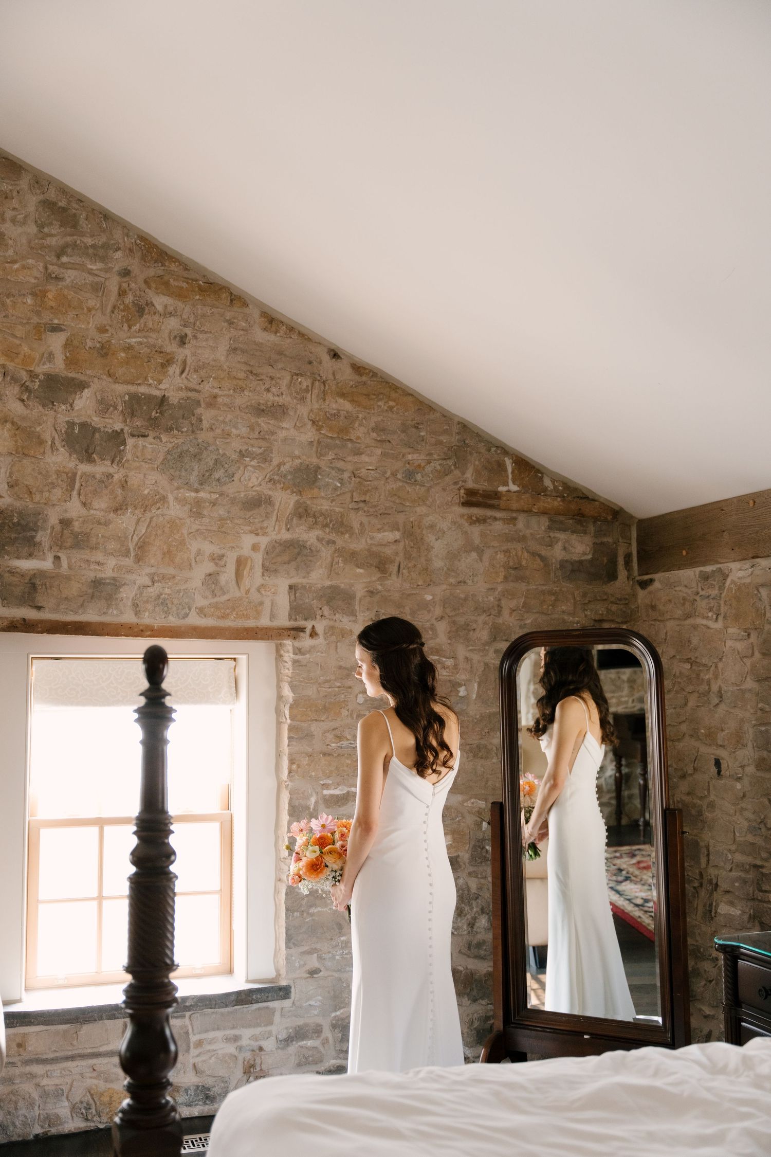 Bride in white gown holding bouquet stands before floor mirror in rustic stone-walled bedroom.