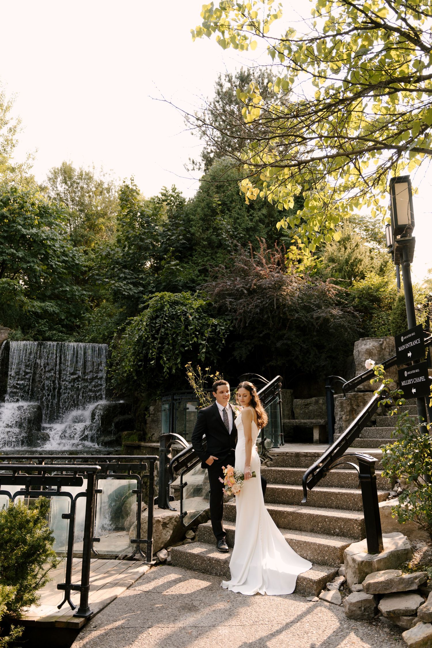 Bride and groom posing on stone steps beside a romantic waterfall in a lush garden Ancaster Mill venue setting.