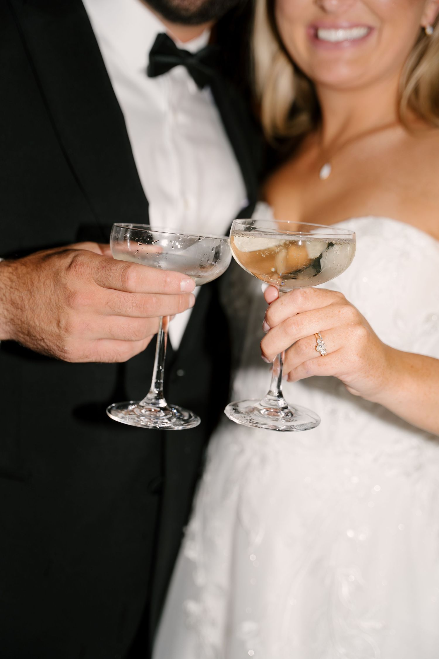 Close-up of bride and groom clinking champagne coupe glasses, showcasing bride's diamond engagement ring.
