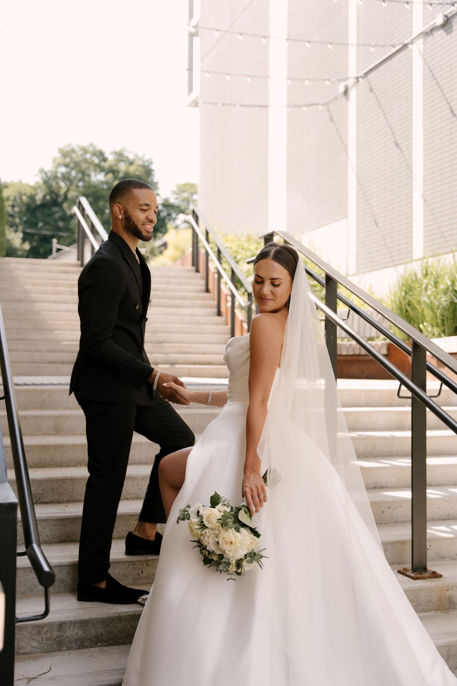 Groom helps bride descend outdoor stairs as she holds a white floral bouquet on their wedding day.