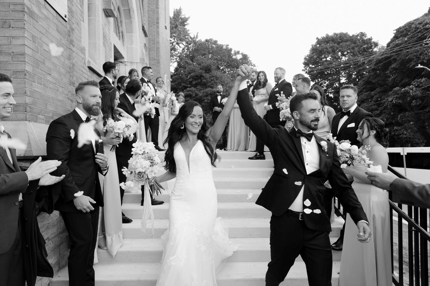 Black and white photo of joyful bride and groom celebrating on church steps with wedding party.