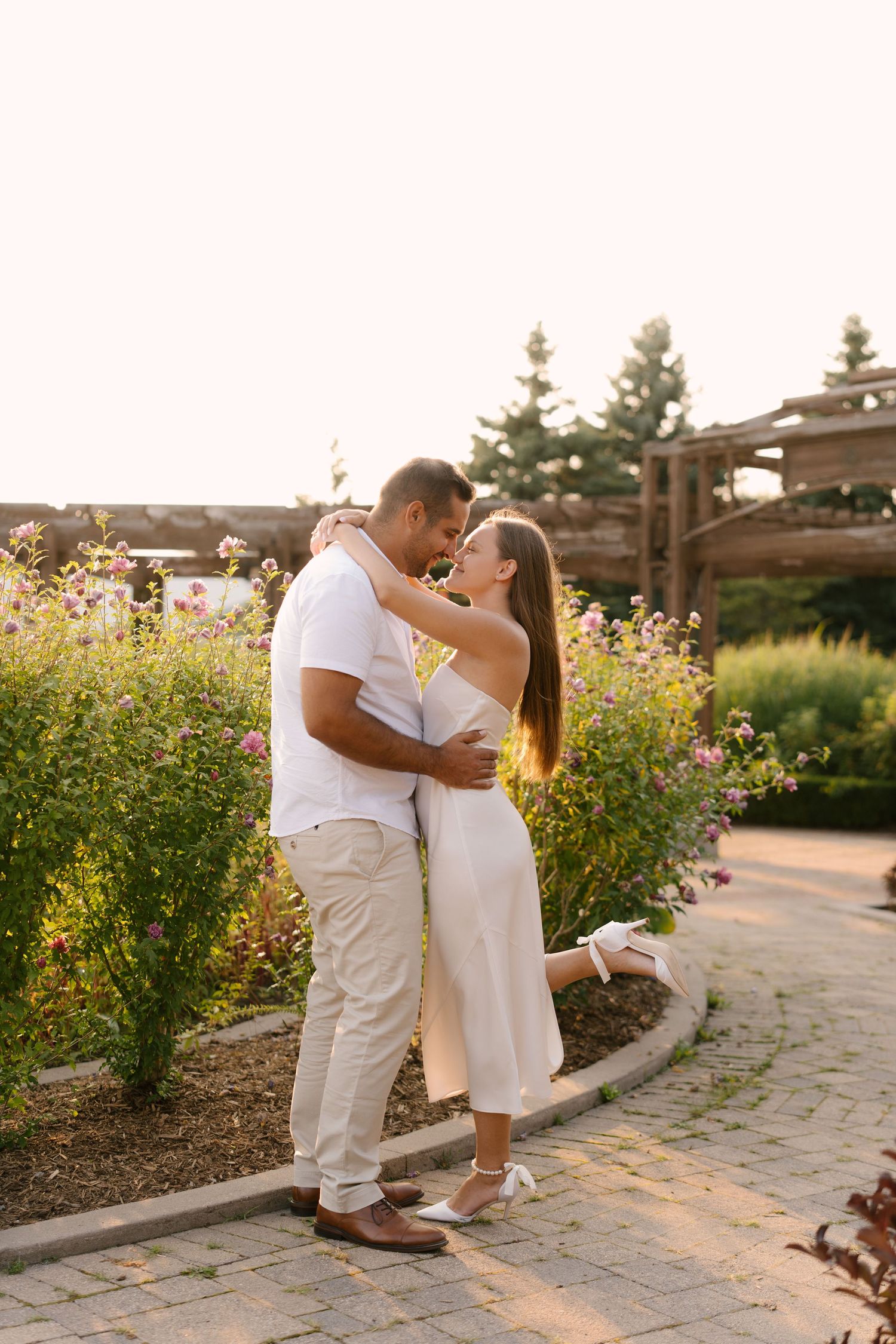 Couple sharing a tender embrace in a vibrant flower garden at golden hour sunset, woman in white dress.