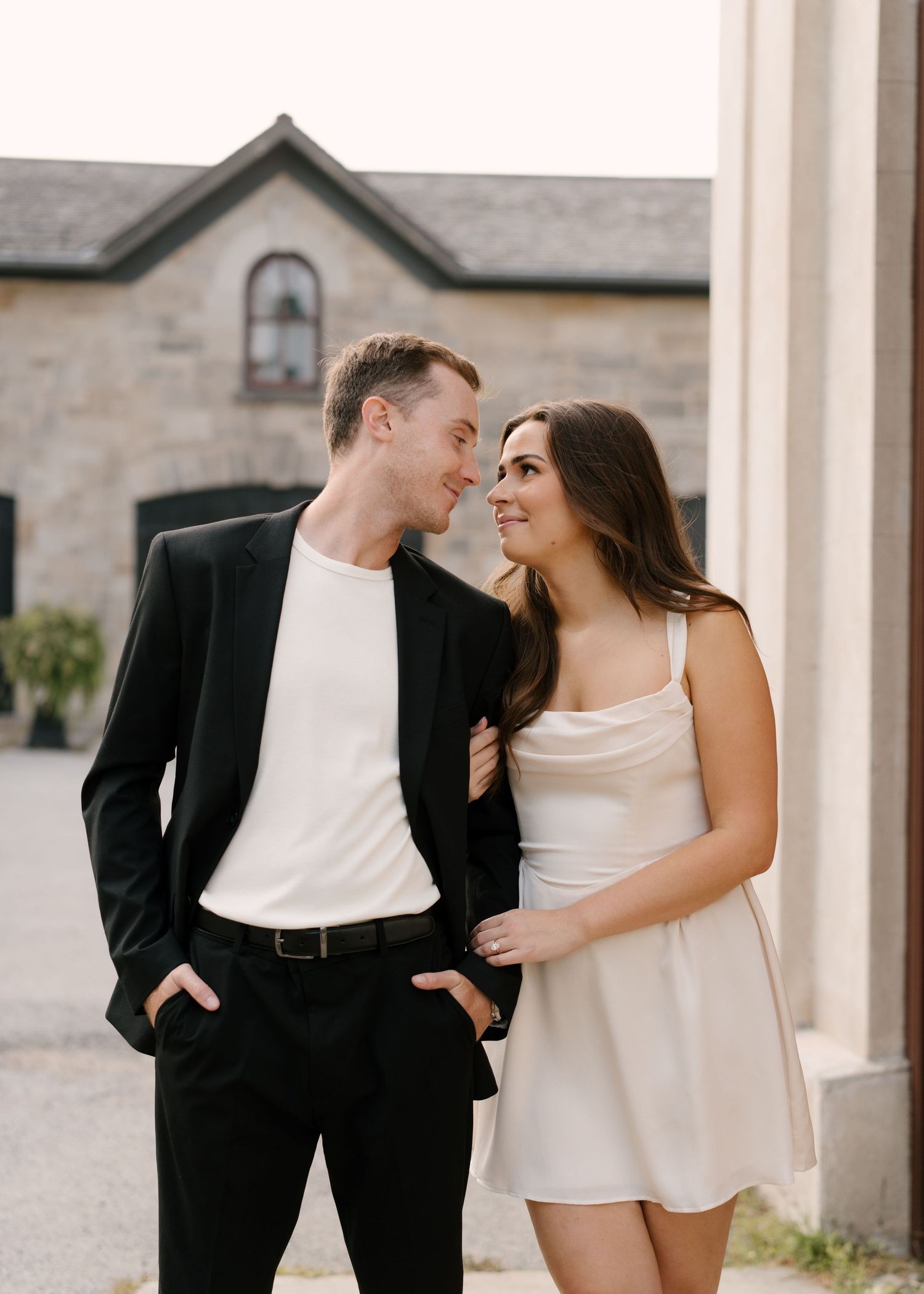 Smiling couple gazing at each other outdoors, man in black blazer and white tee, woman in cream mini dress.