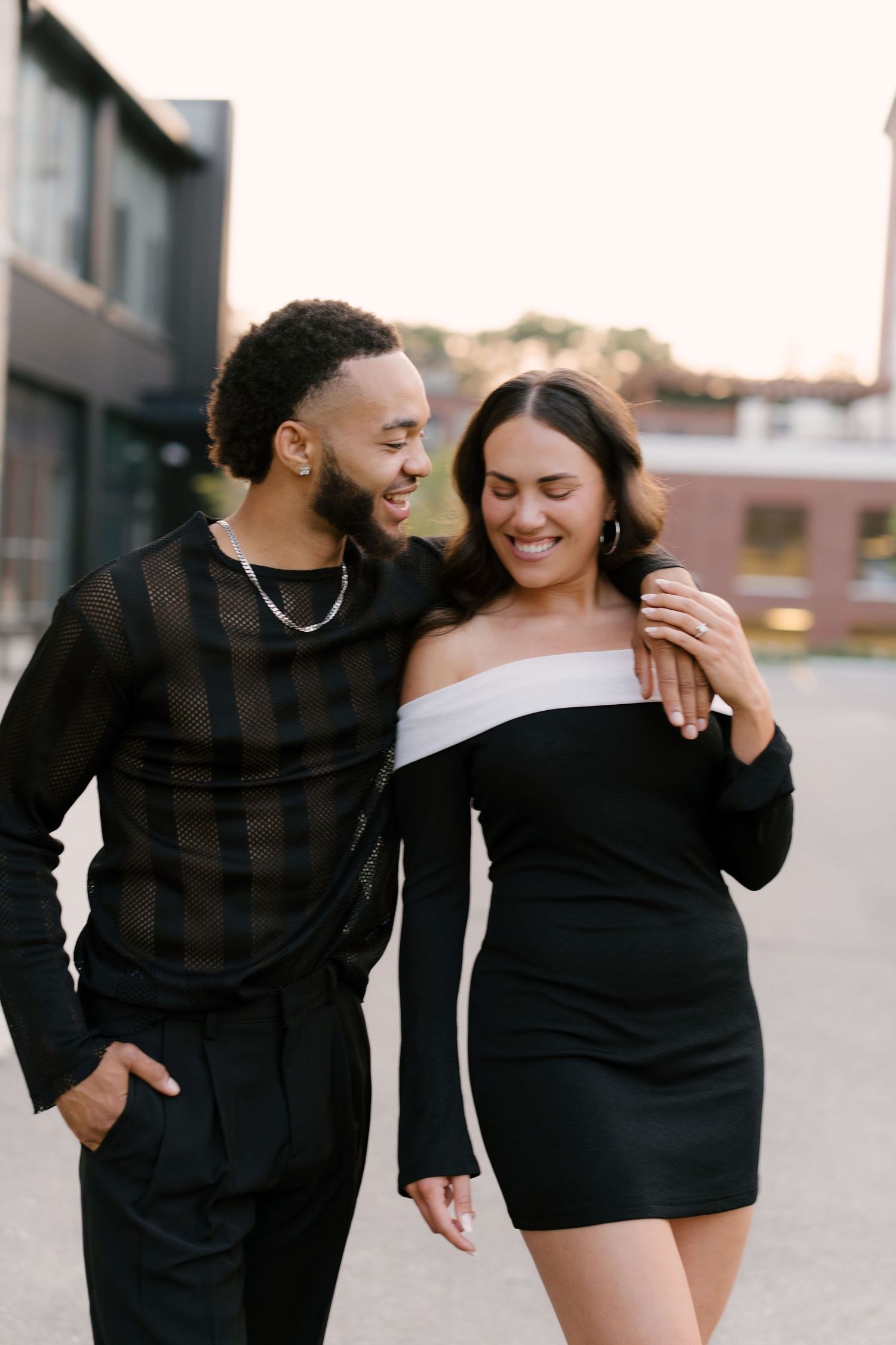 Smiling couple in matching black outfits walking together outdoors, man with arm around woman's shoulders.