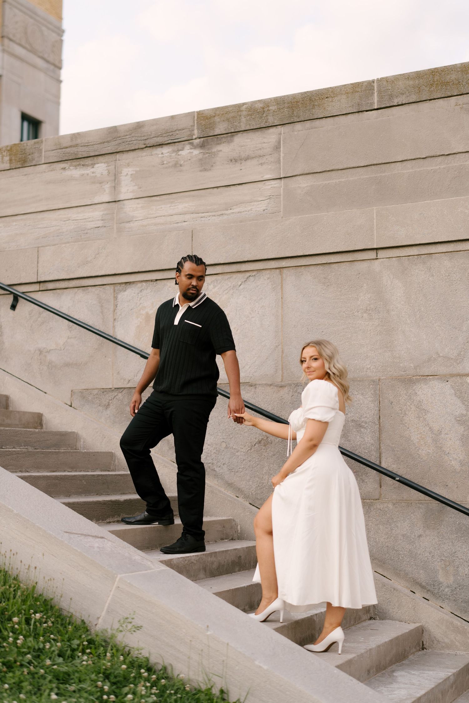 An interracial couple holds hands while posing on stone steps beside a large granite building.