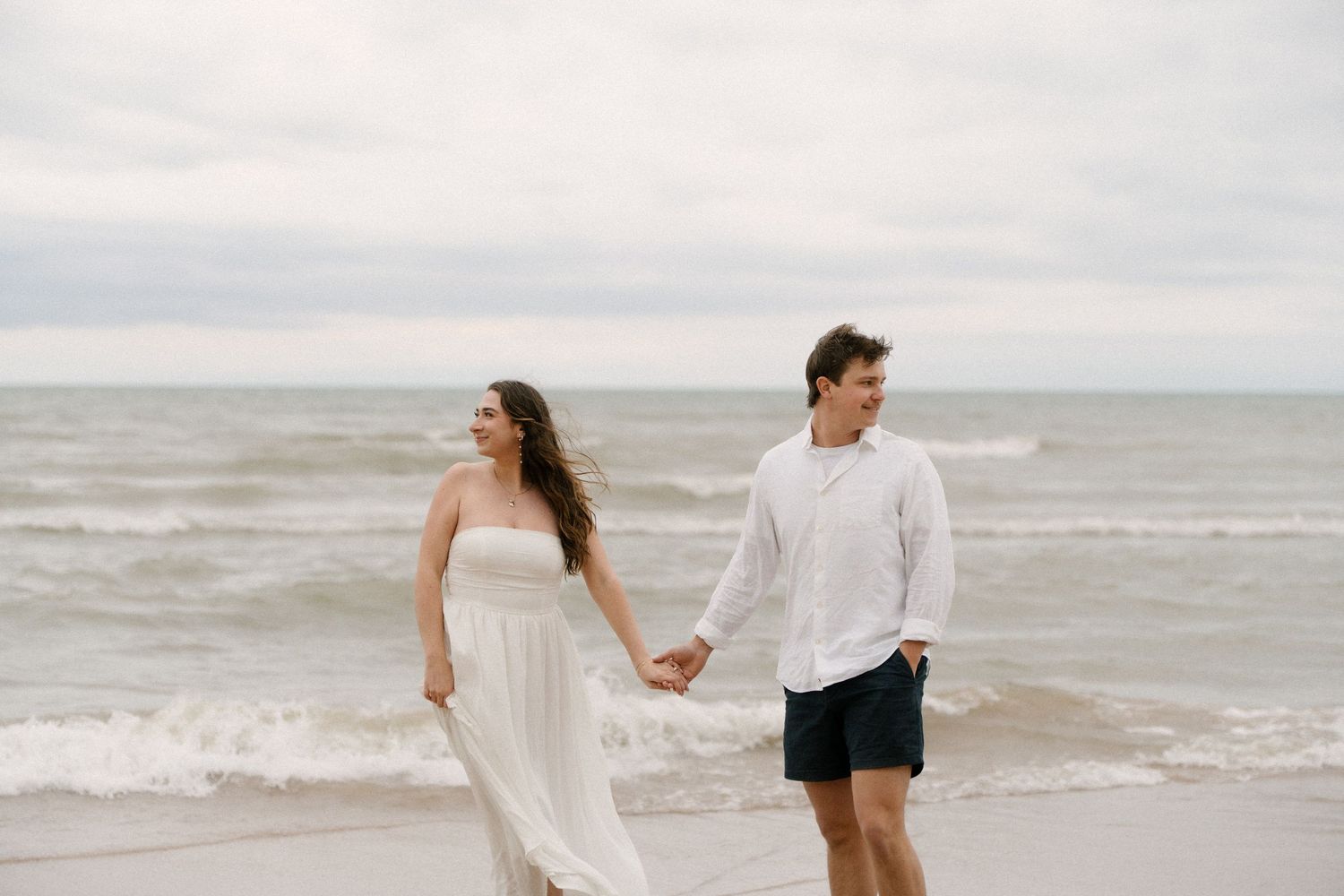 Couple holding hands walking along sandy beach, woman in white strapless dress, man in white shirt and navy shorts.