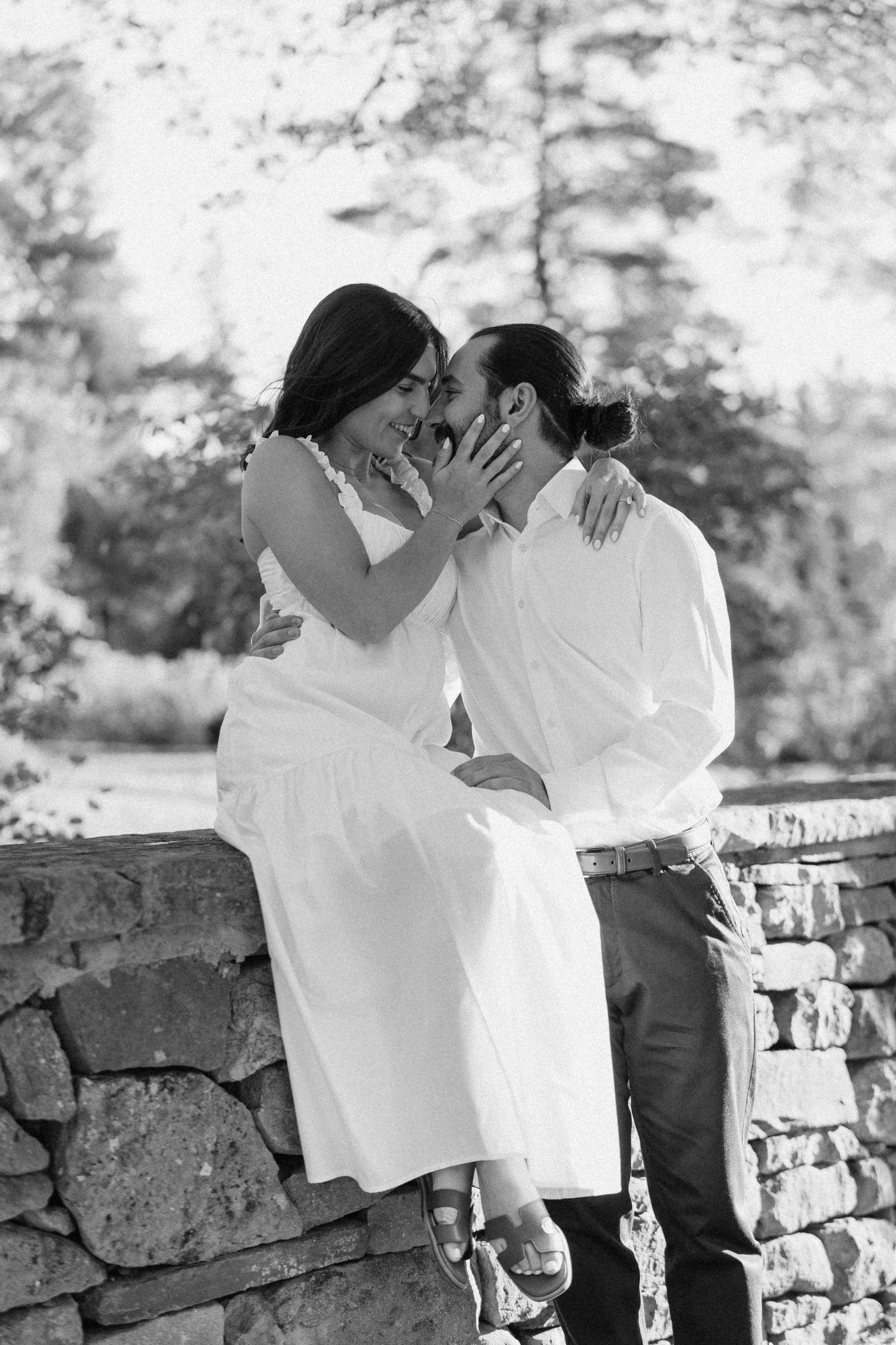 Black and white photo of smiling couple sharing a tender kiss on a rustic stone wall.