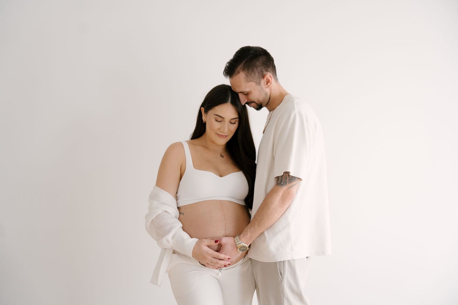 Couple in neutral tones tenderly cradling pregnant belly together against white background in maternity portrait.