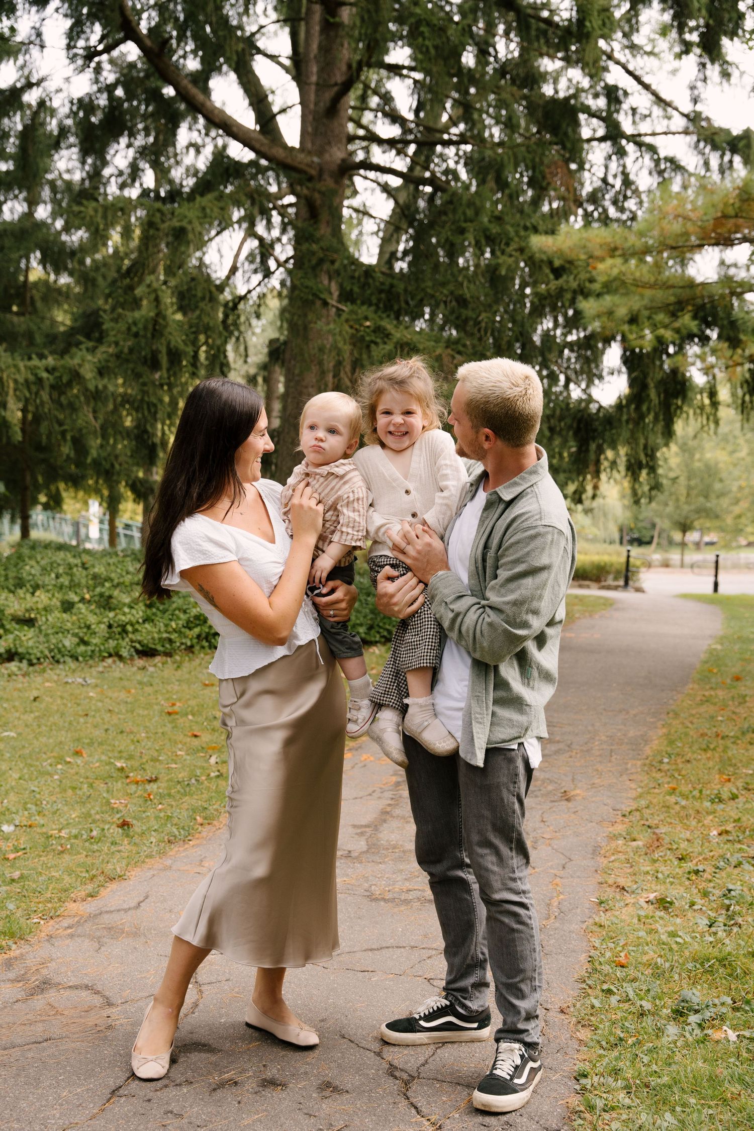 Parents holding two young children on park path, smiling and interacting outdoors.