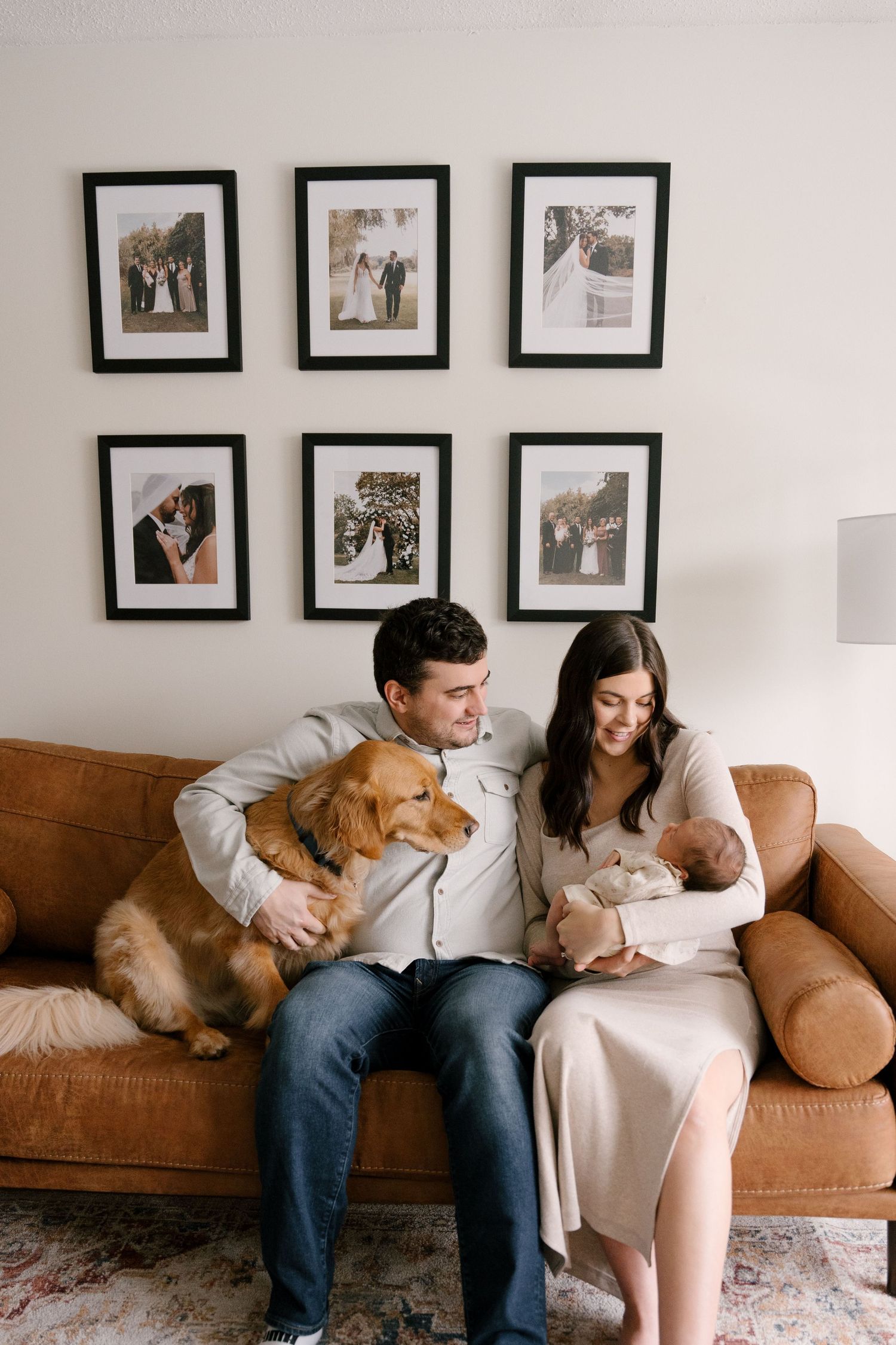 Couple with newborn and golden retriever sitting on leather sofa beneath framed wedding photos.
