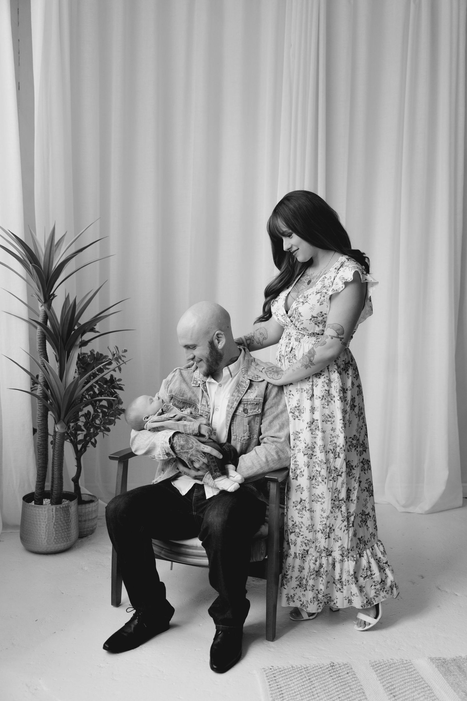 Black and white photo of a bald father sitting with newborn while mother in floral dress stands beside.