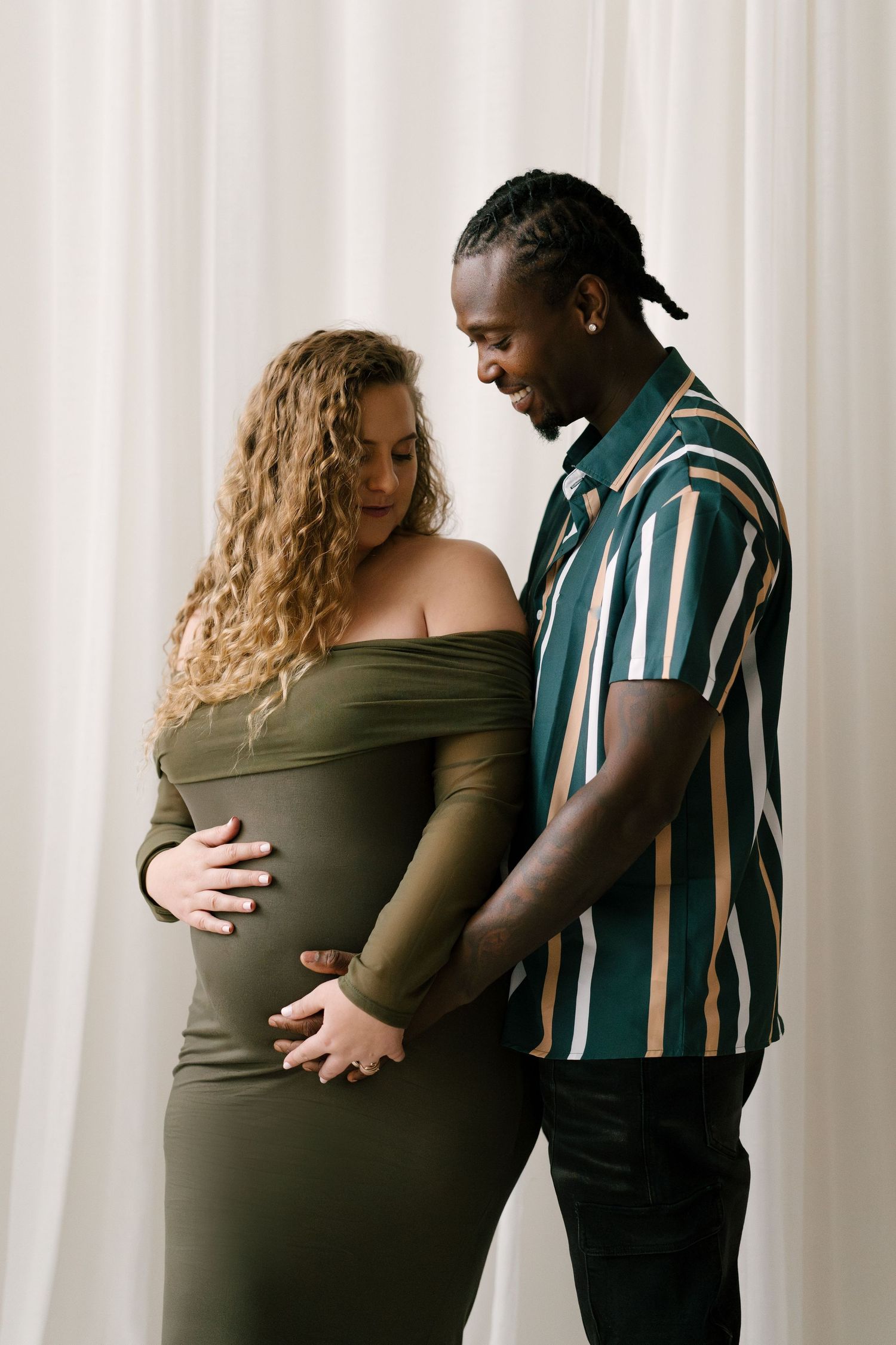 Smiling interracial couple embracing pregnant belly together against soft white curtain backdrop.