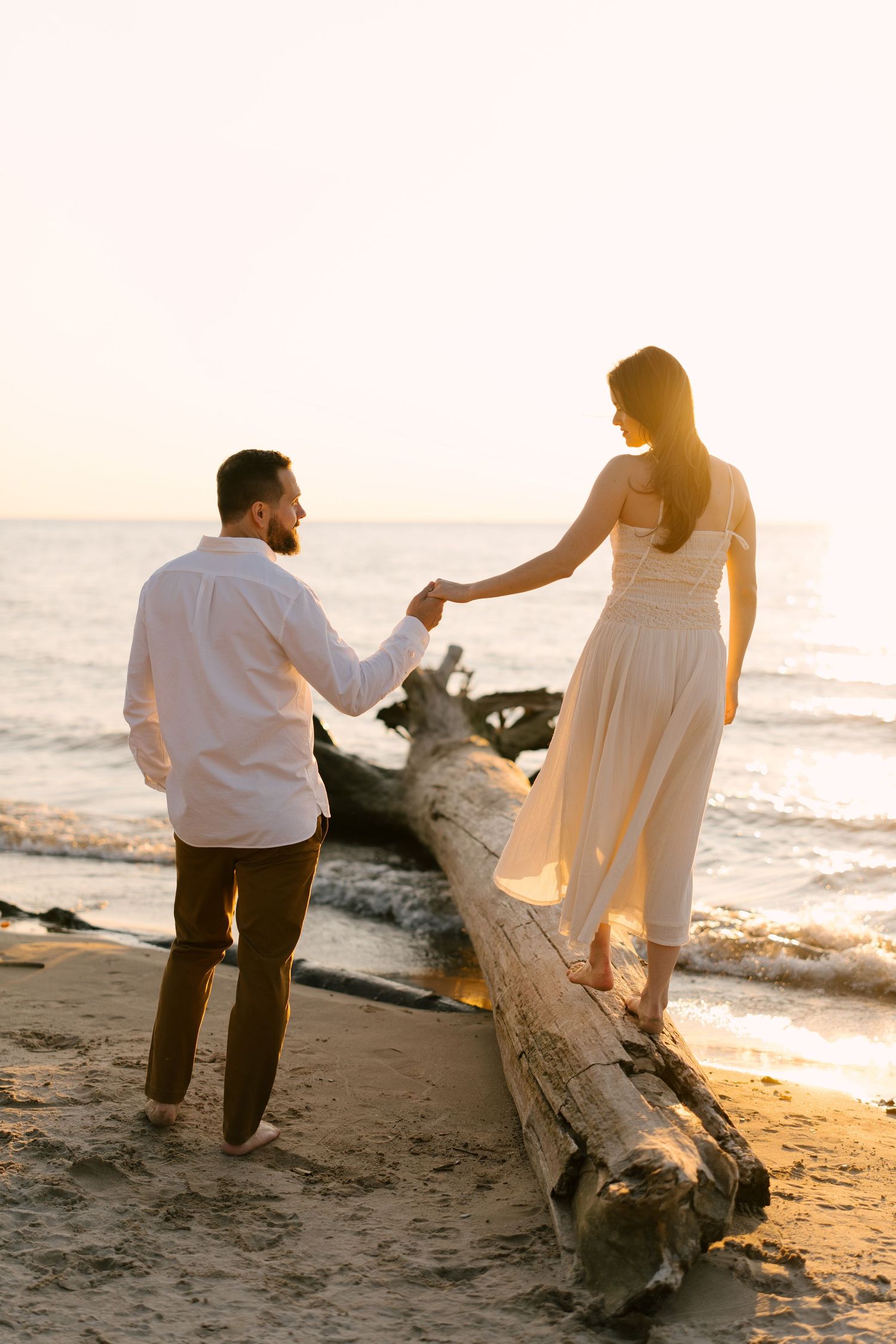 Couple holding hands on a driftwood log at a beach during sunset, woman in white dress balancing gracefully.
