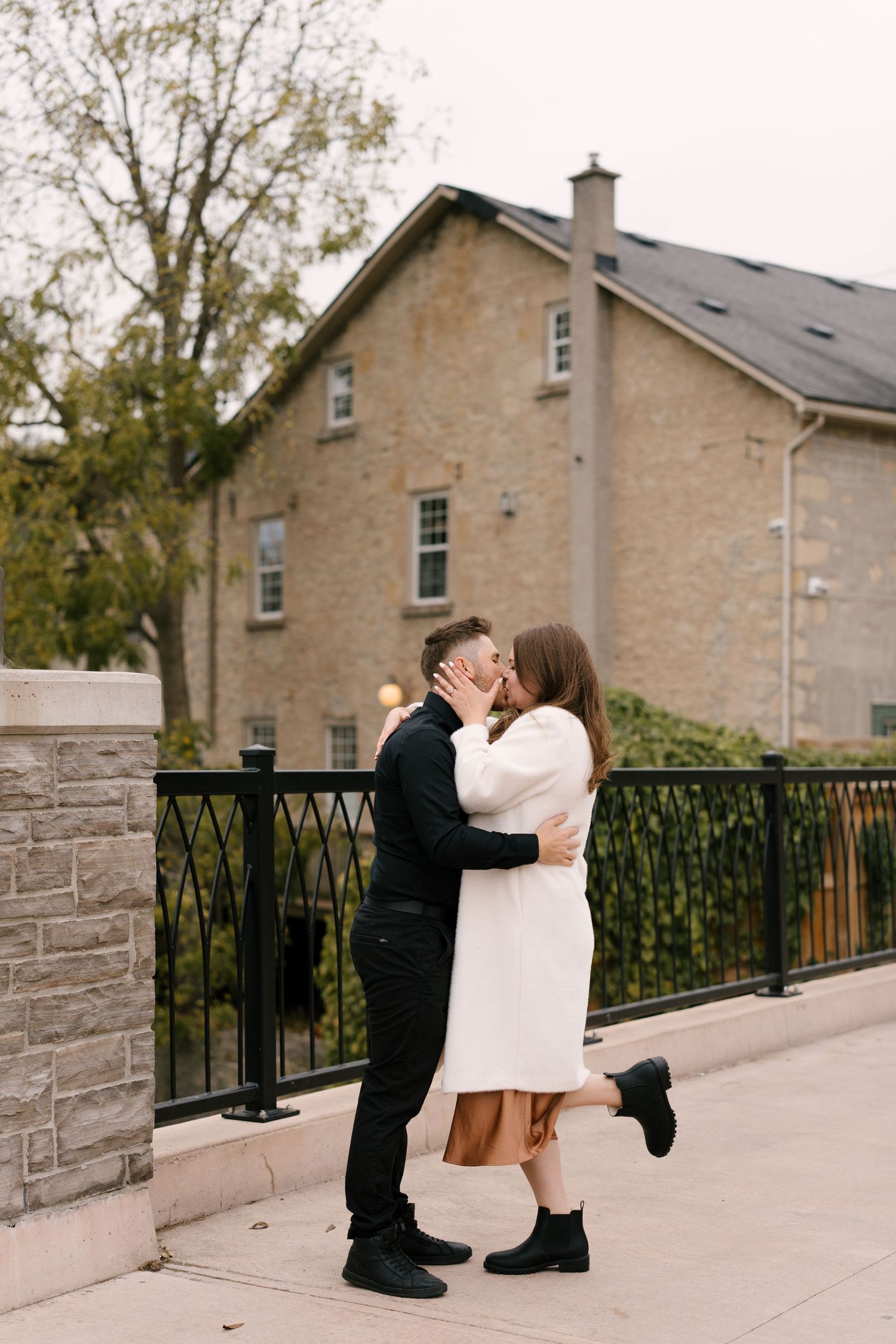 Couple sharing a romantic kiss on a bridge walkway with a historic stone building in the background during fall.