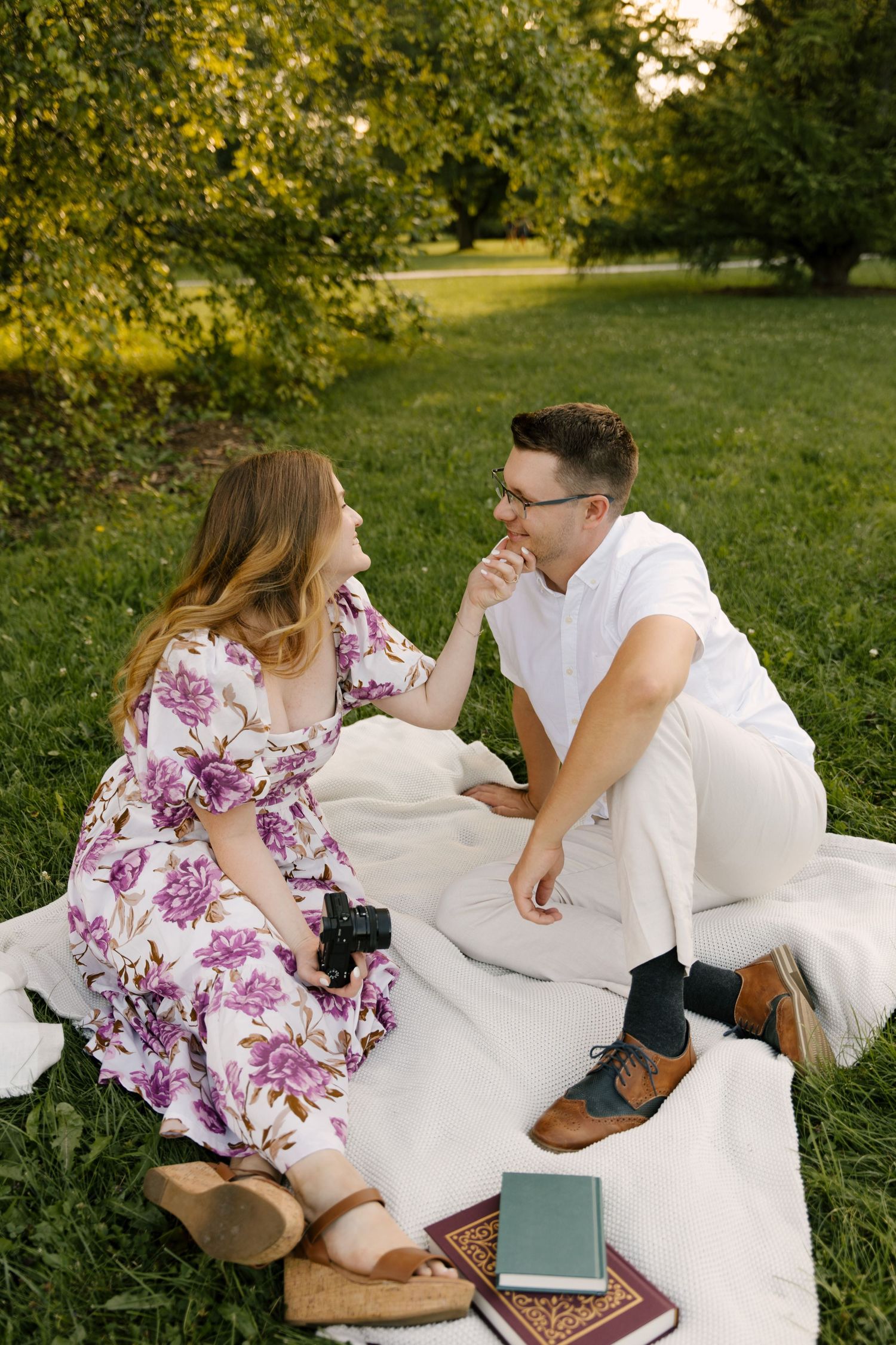 Couple sitting on a white picnic blanket in a park, smiling at each other with books nearby.