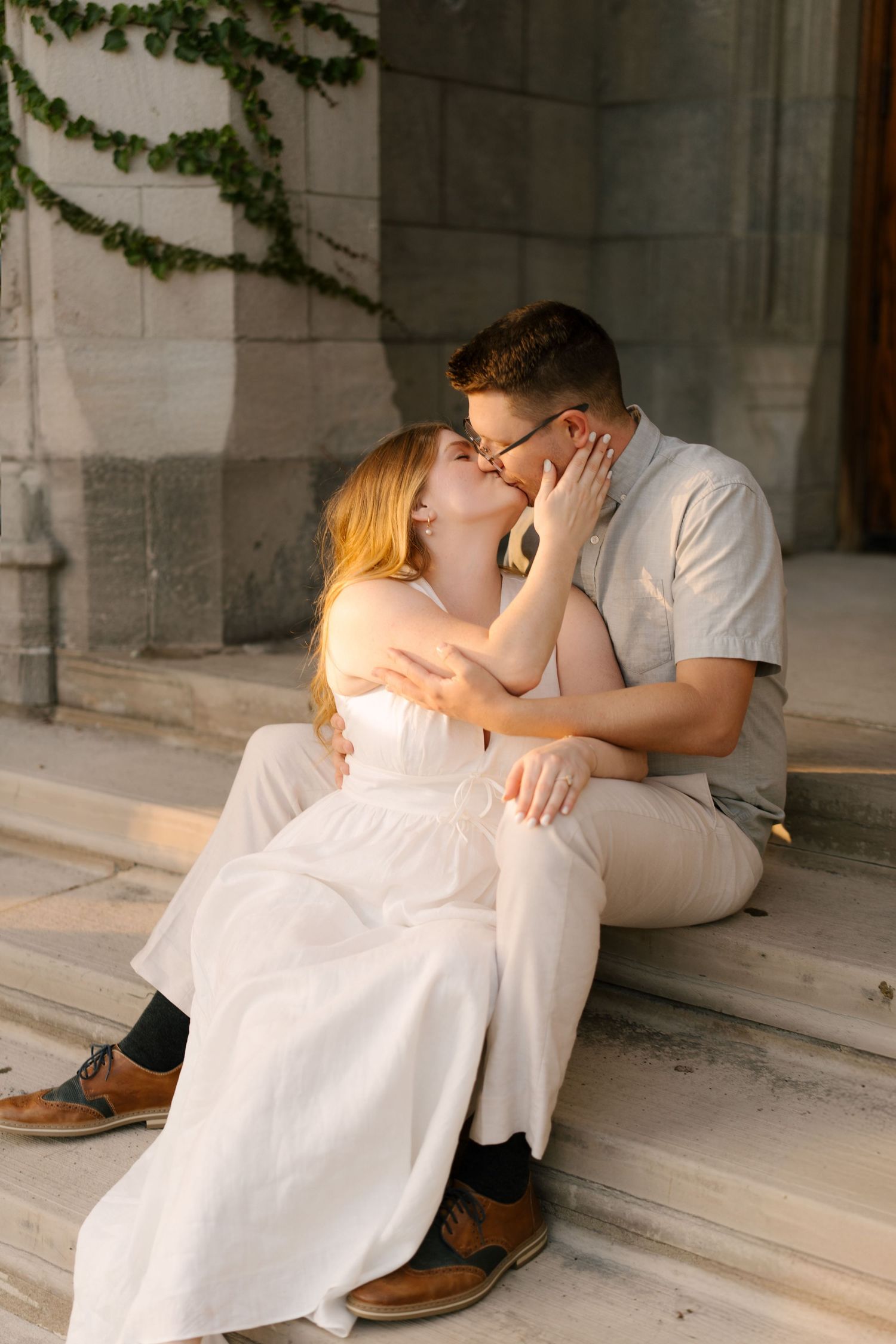 Romantic couple sharing a kiss on stone steps, woman in white dress, warm sunset light.