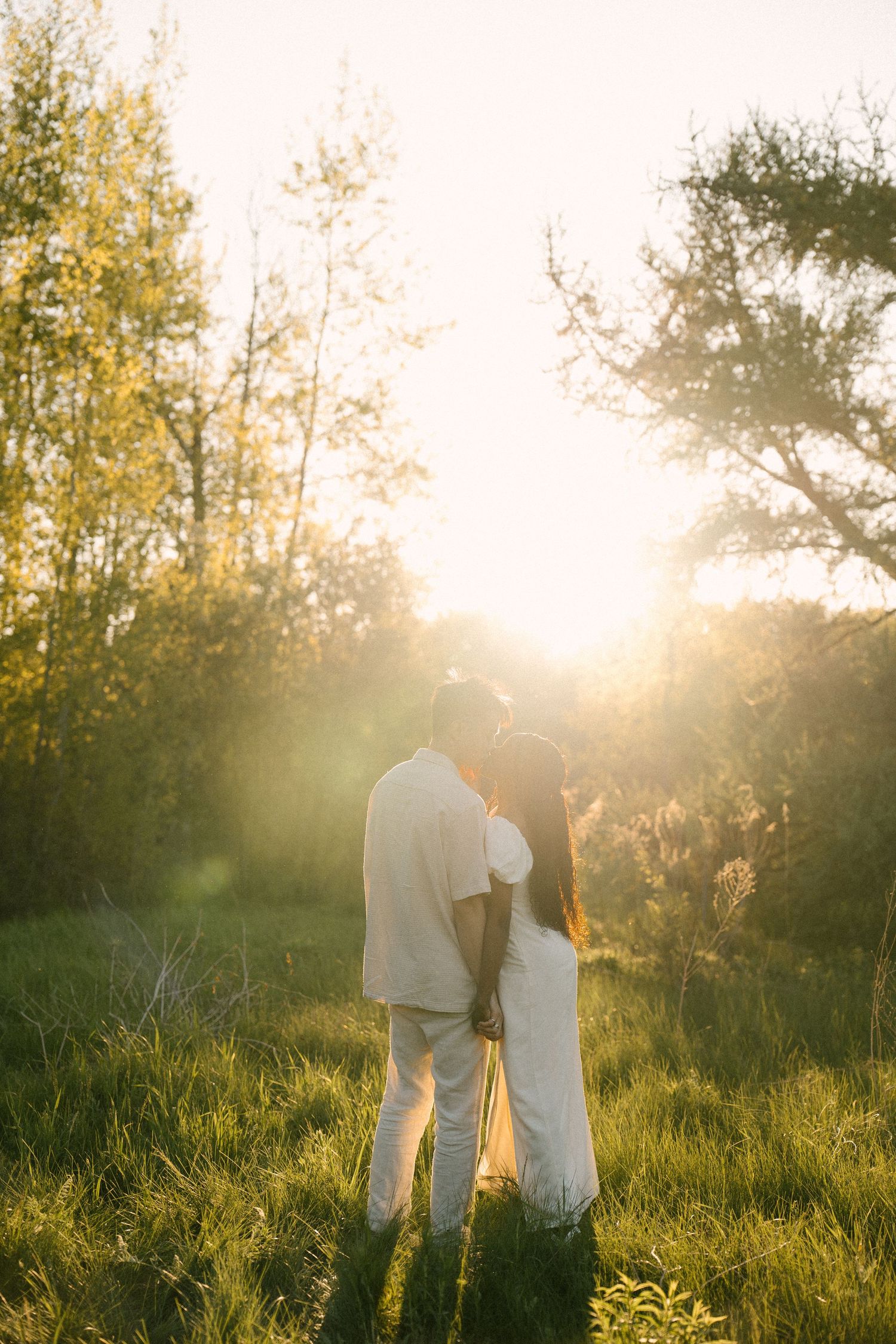 Couple holding hands in golden sunset light among lush green trees in a meadow.