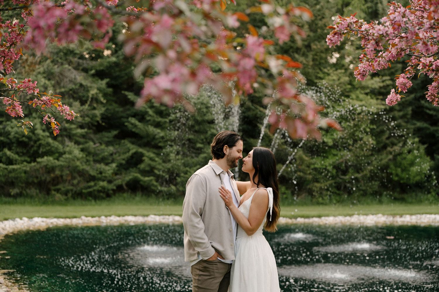 Romantic couple almost kissing near a garden fountain framed by pink blossoming tree branches.