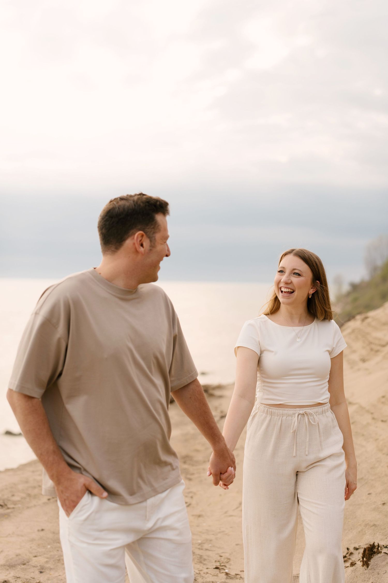 Happy couple holding hands and laughing while walking on sandy beach at dusk.