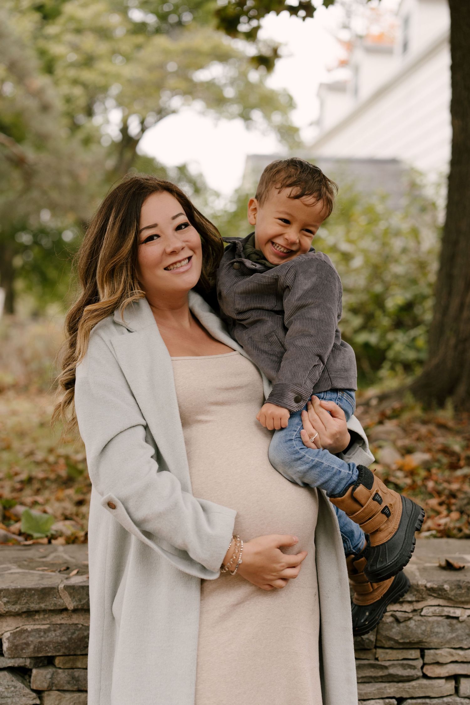 Smiling pregnant woman in cream dress and grey coat holding happy toddler boy outdoors.