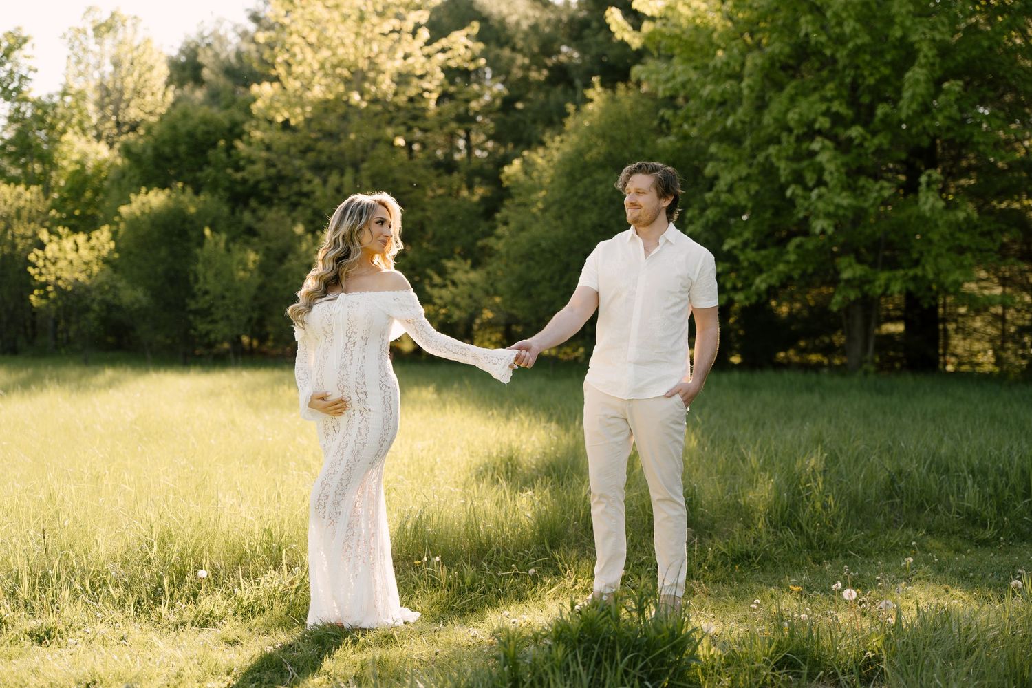Expecting couple holding hands and smiling in a sunny green meadow during outdoor maternity photoshoot.