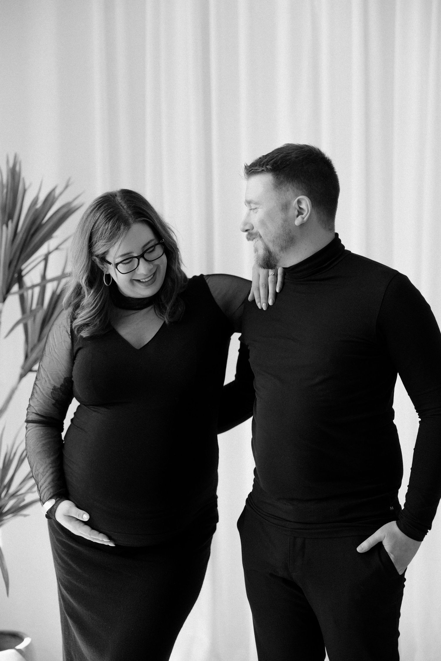 Black and white photo of smiling pregnant woman and partner in black outfits standing together indoors.