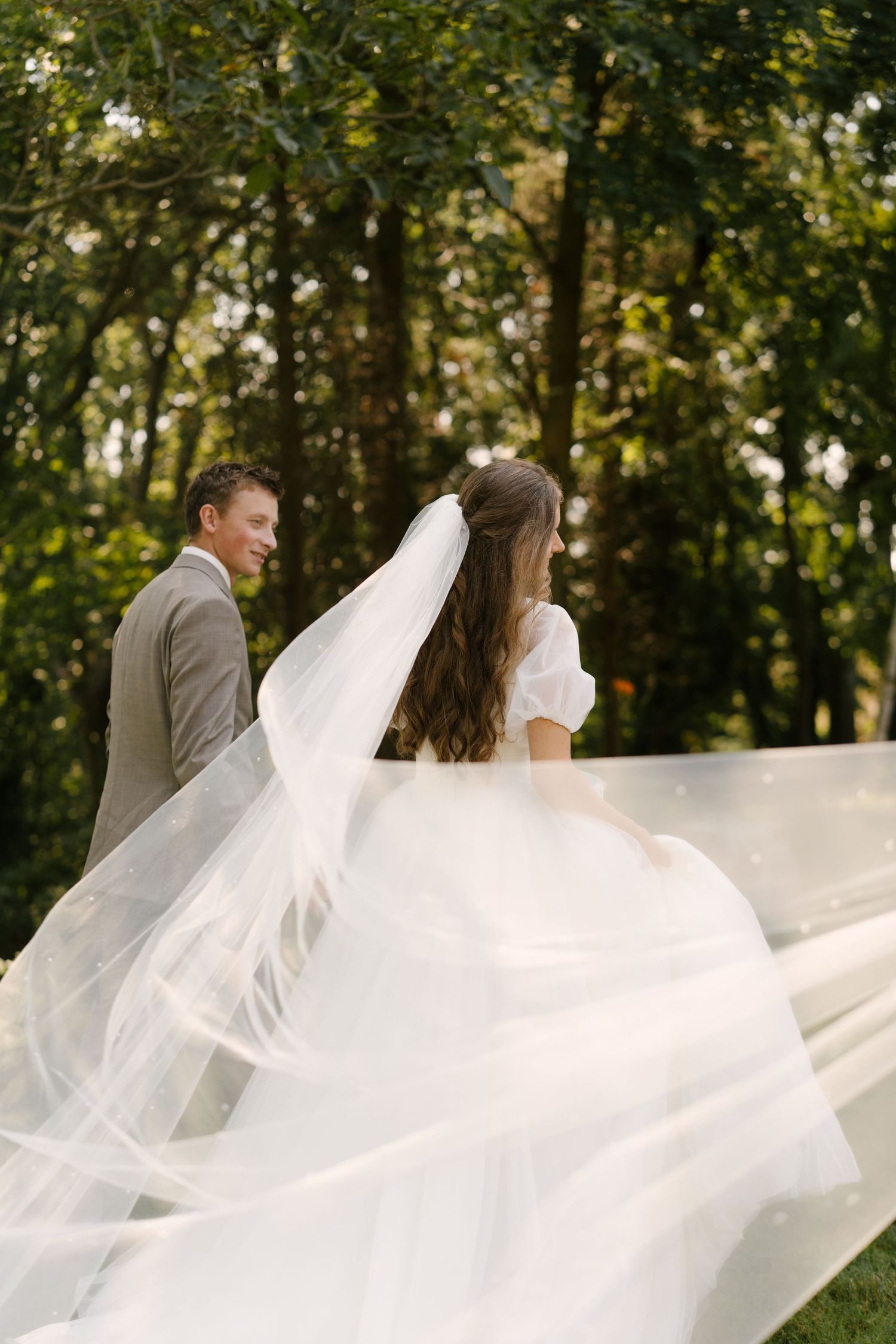 Bride and groom walking in forest as flowing white veil billows dramatically in the wind.