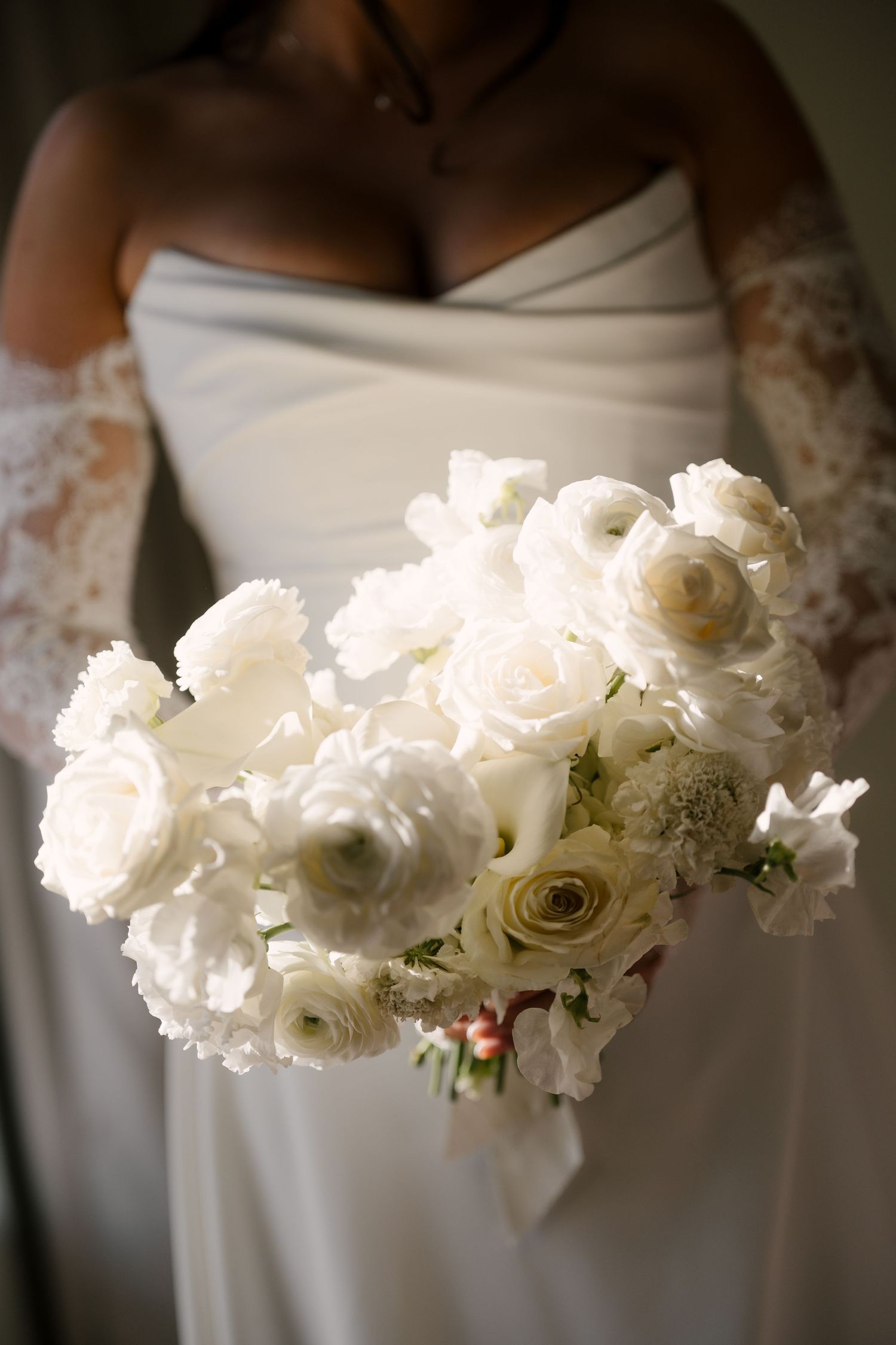 Bride in strapless white gown holds lush all-white bouquet of ranunculus and roses with lace gloves.
