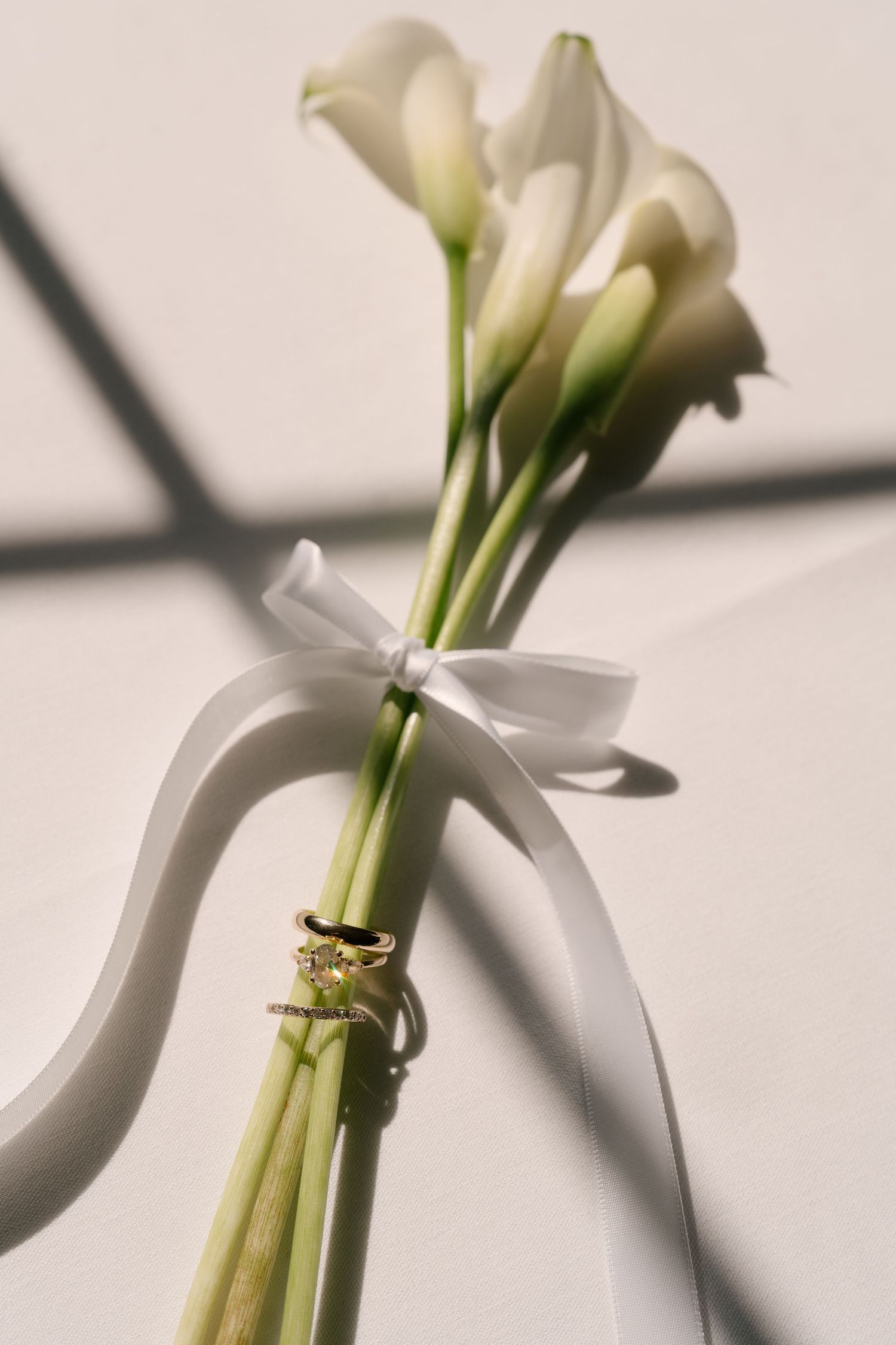 Elegant calla lily bouquet with white satin bow and gold rings in natural sunlight.