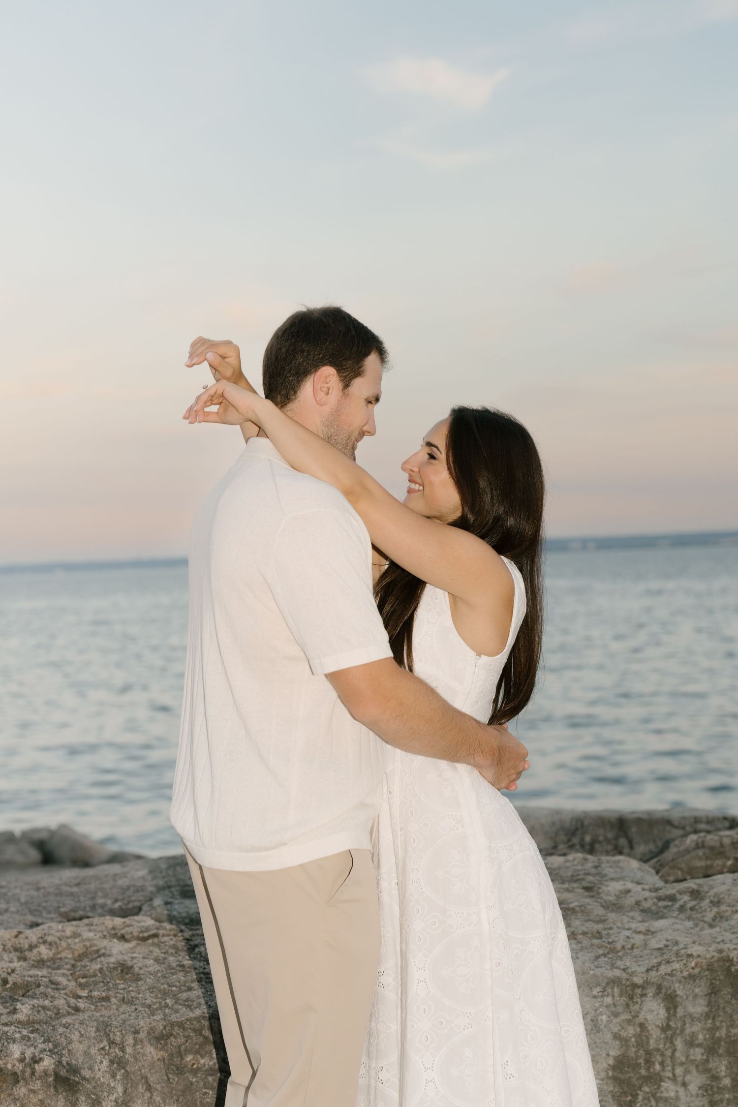 Couple sharing tender embrace by rocky shoreline at dusk, woman in white dress smiling at partner.