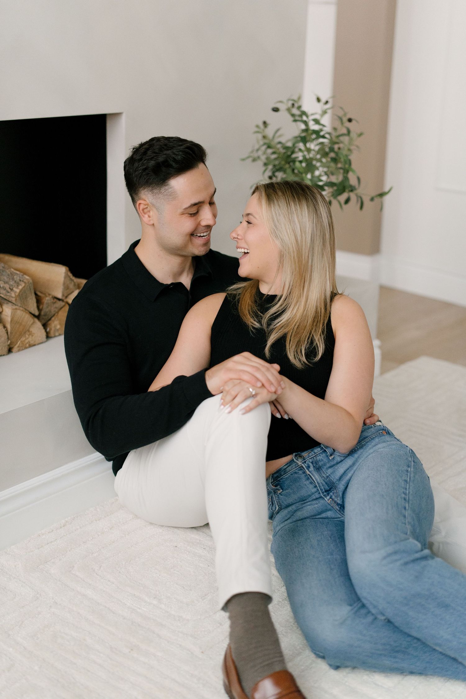 Smiling couple sitting on white rug near cozy fireplace.